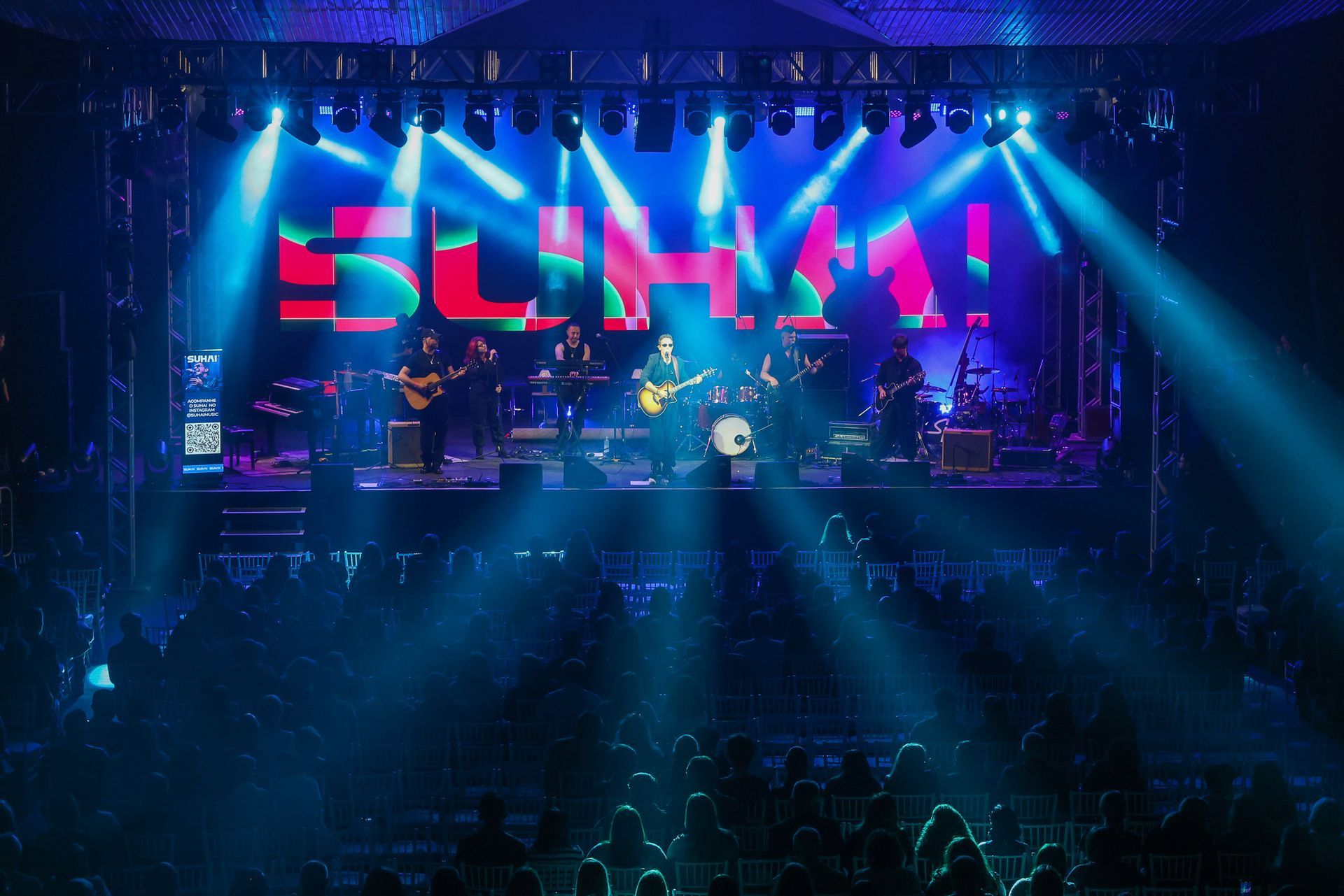 Uma banda se apresenta em um palco bem iluminado por holofotes azuis, com a palavra 