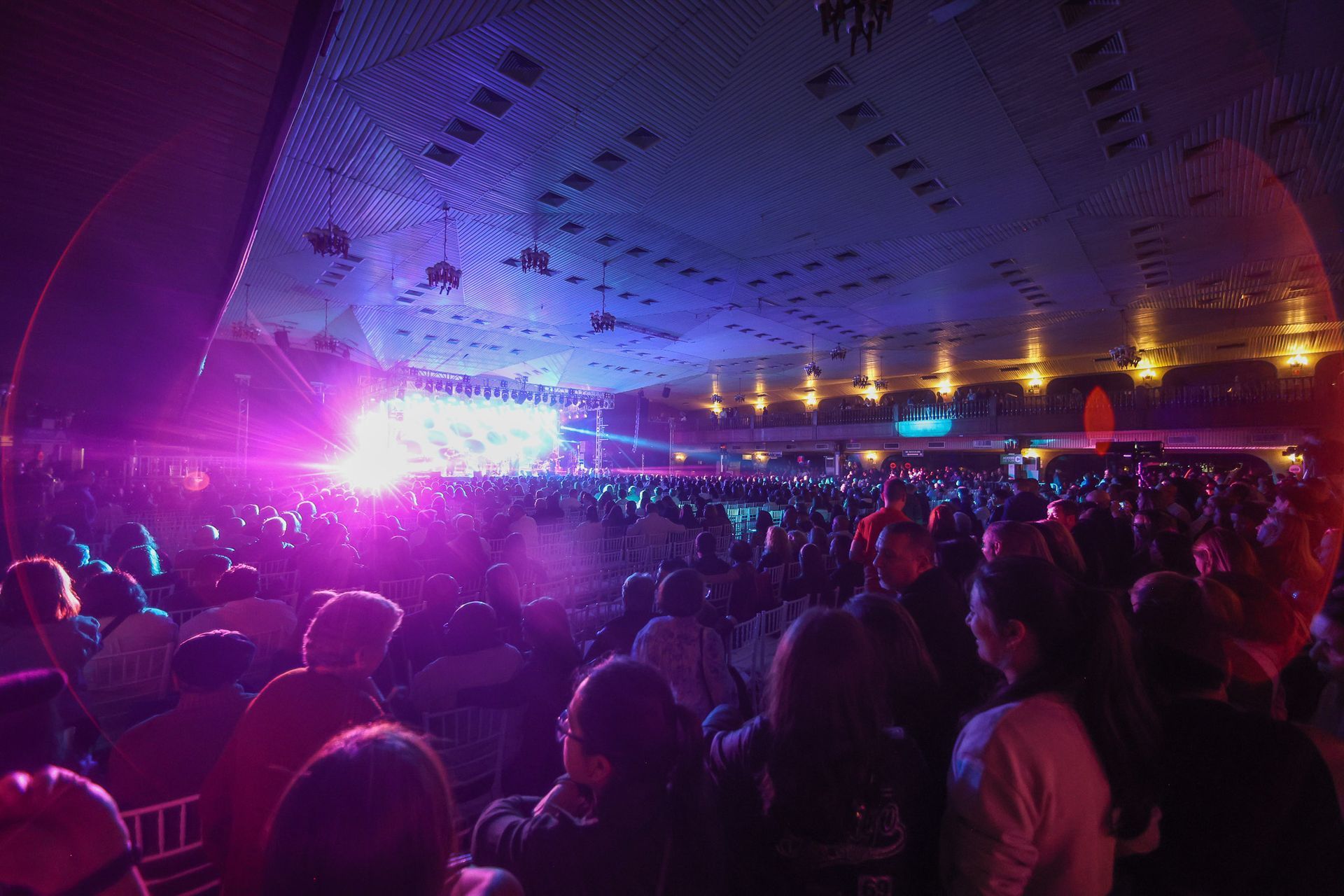 Uma multidão de pessoas  e com pouca luz, de frente para um palco brilhantemente iluminado com luzes roxas vibrantes.