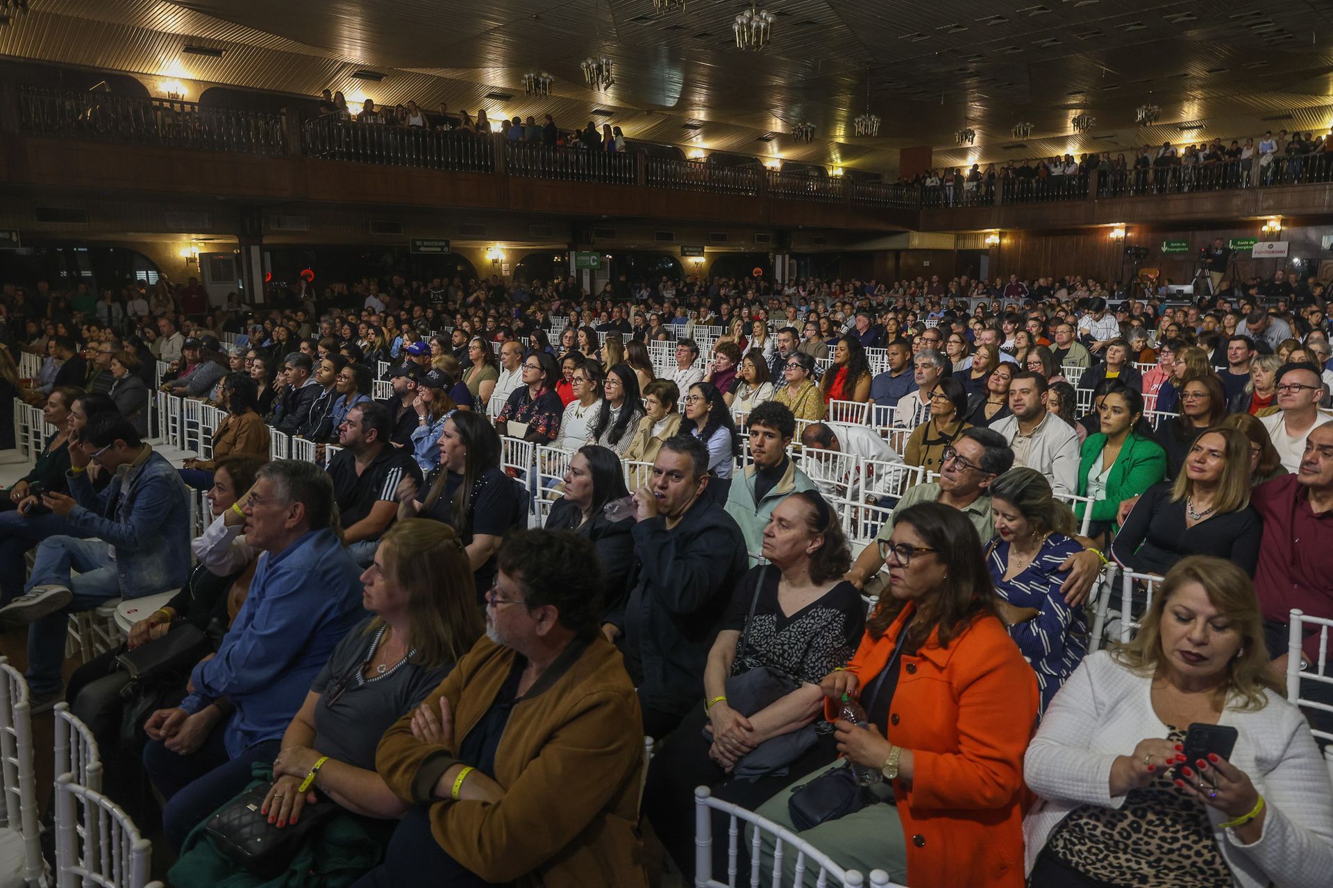 Um grande auditório, com iluminação tênue, repleto de pessoas sentadas assistindo a um evento.