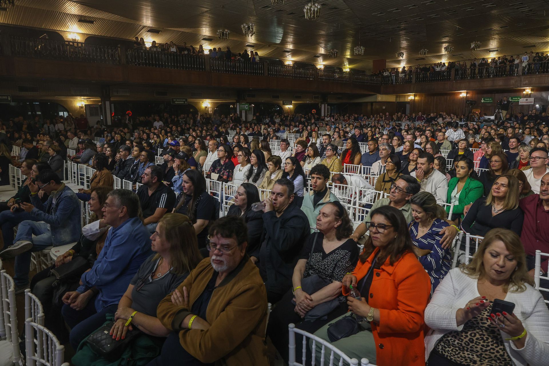 Um público numeroso e diversificado, sentado em um local fechado, para assistir a uma apresentação ou performance.