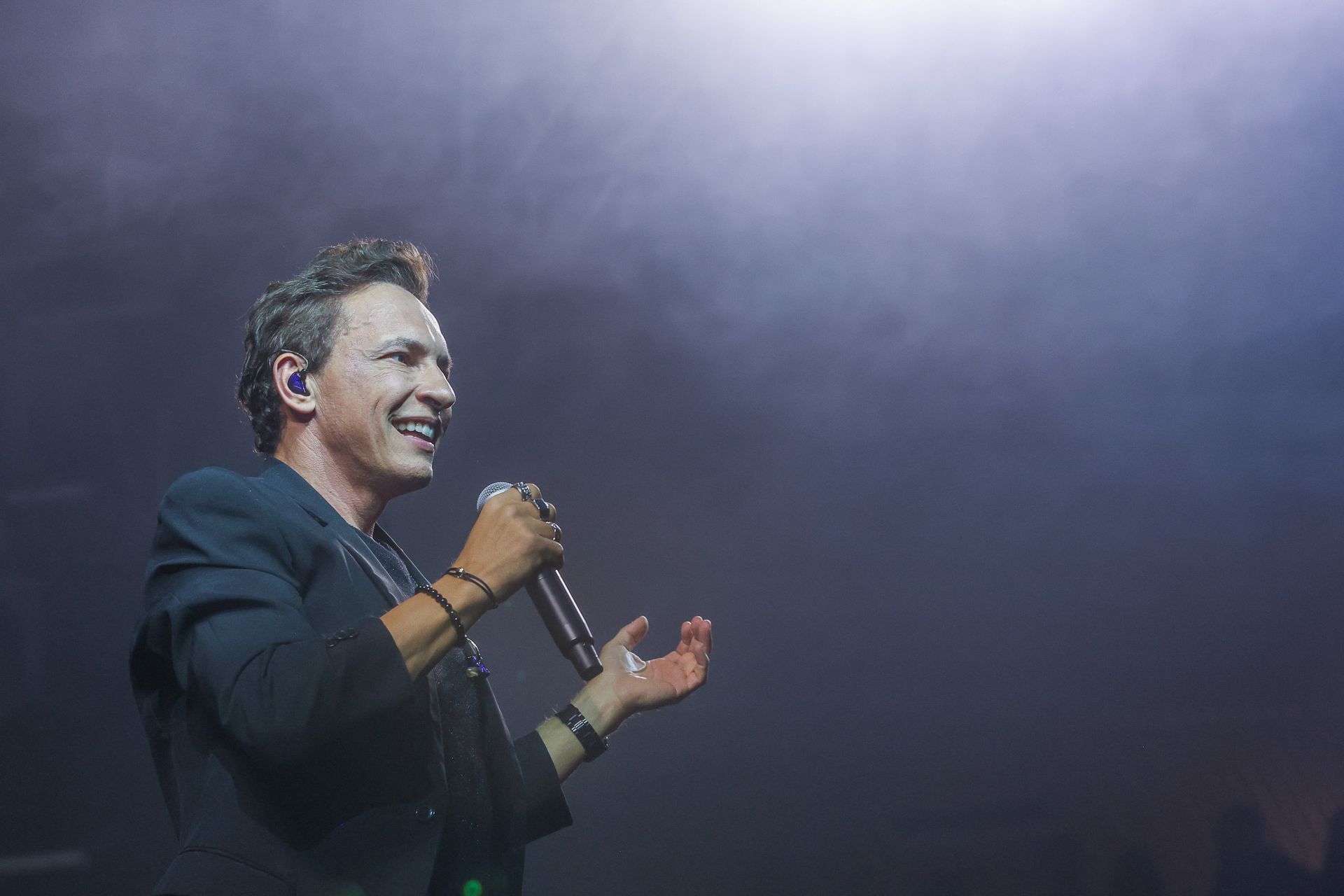 Um artista em um palco sob iluminação suave sorri enquanto fala ao microfone, gesticulando com a mão aberta.