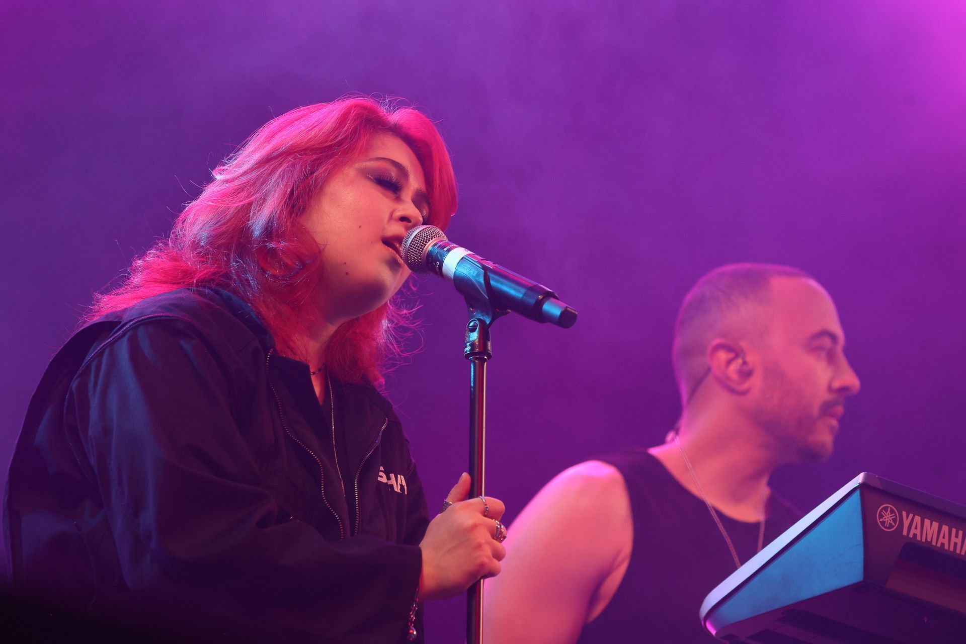 Uma vocalista ruiva canta em um microfone no palco ao lado de um tecladista, iluminados por luzes de palco roxas.
