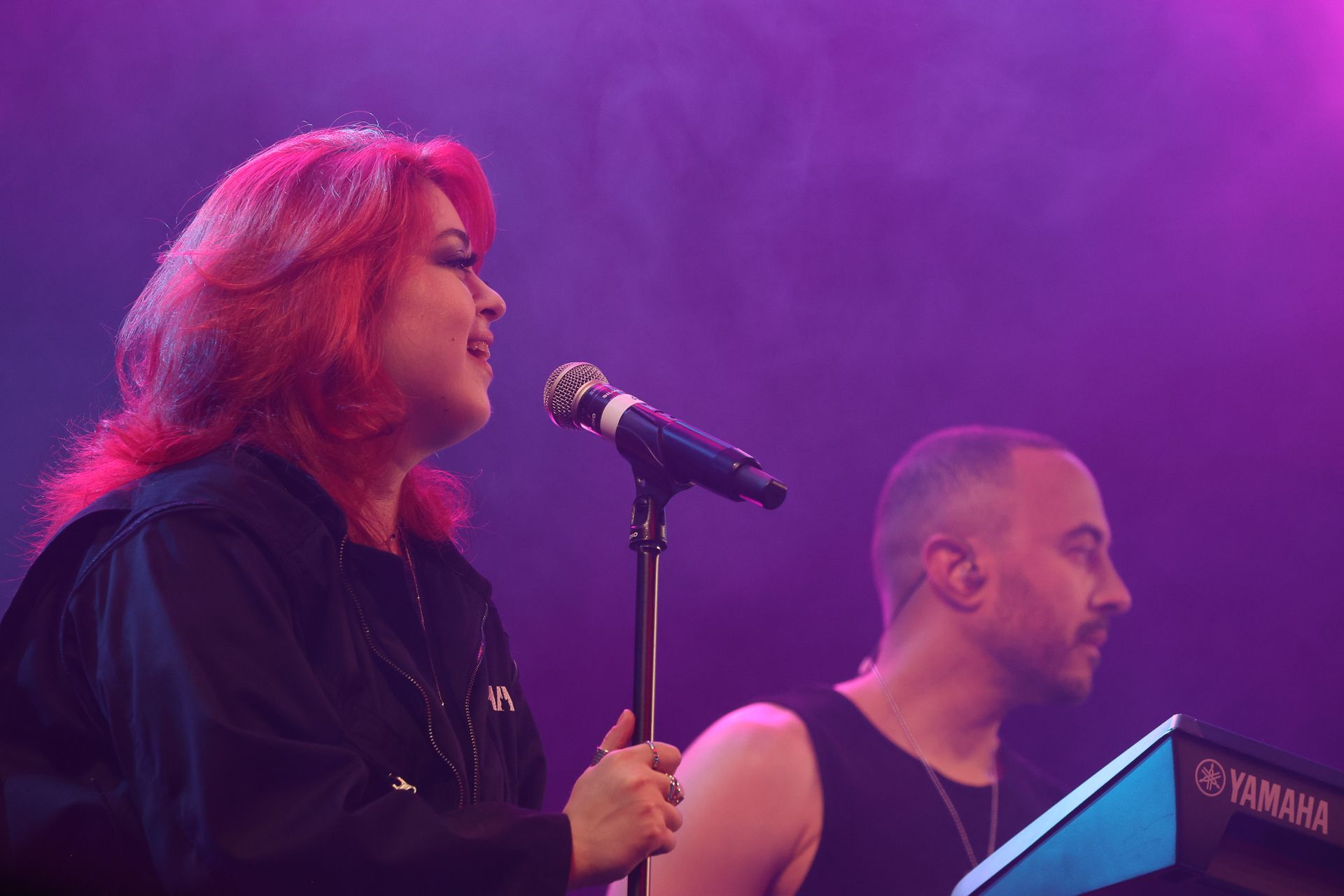 Uma artista ruiva canta em um microfone no palco, acompanhada por um músico que toca teclado sob uma iluminação roxa.