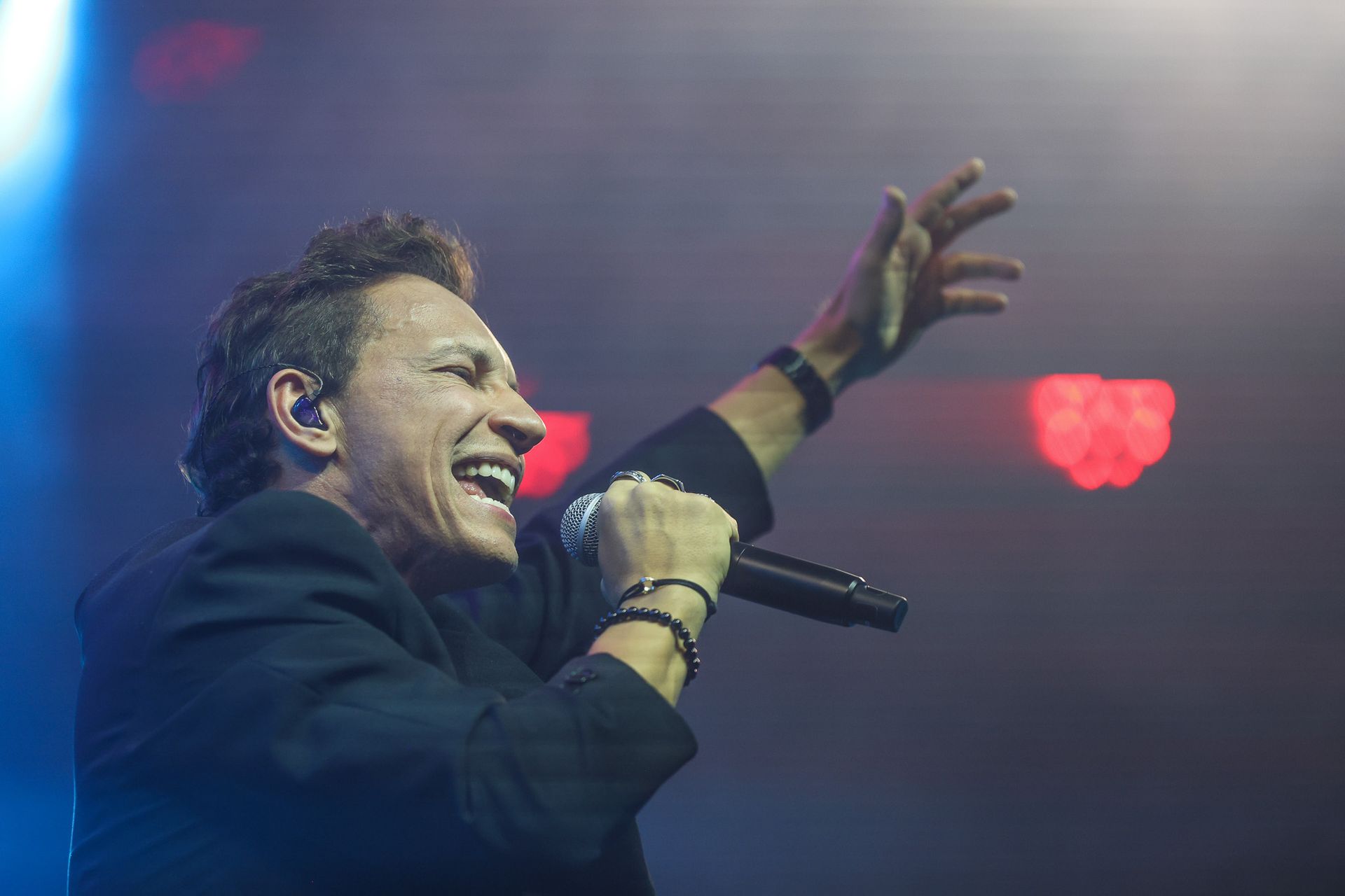 Um cantor no palco segurando um microfone com uma das mãos levantadas, iluminada intensamente por luzes de palco .