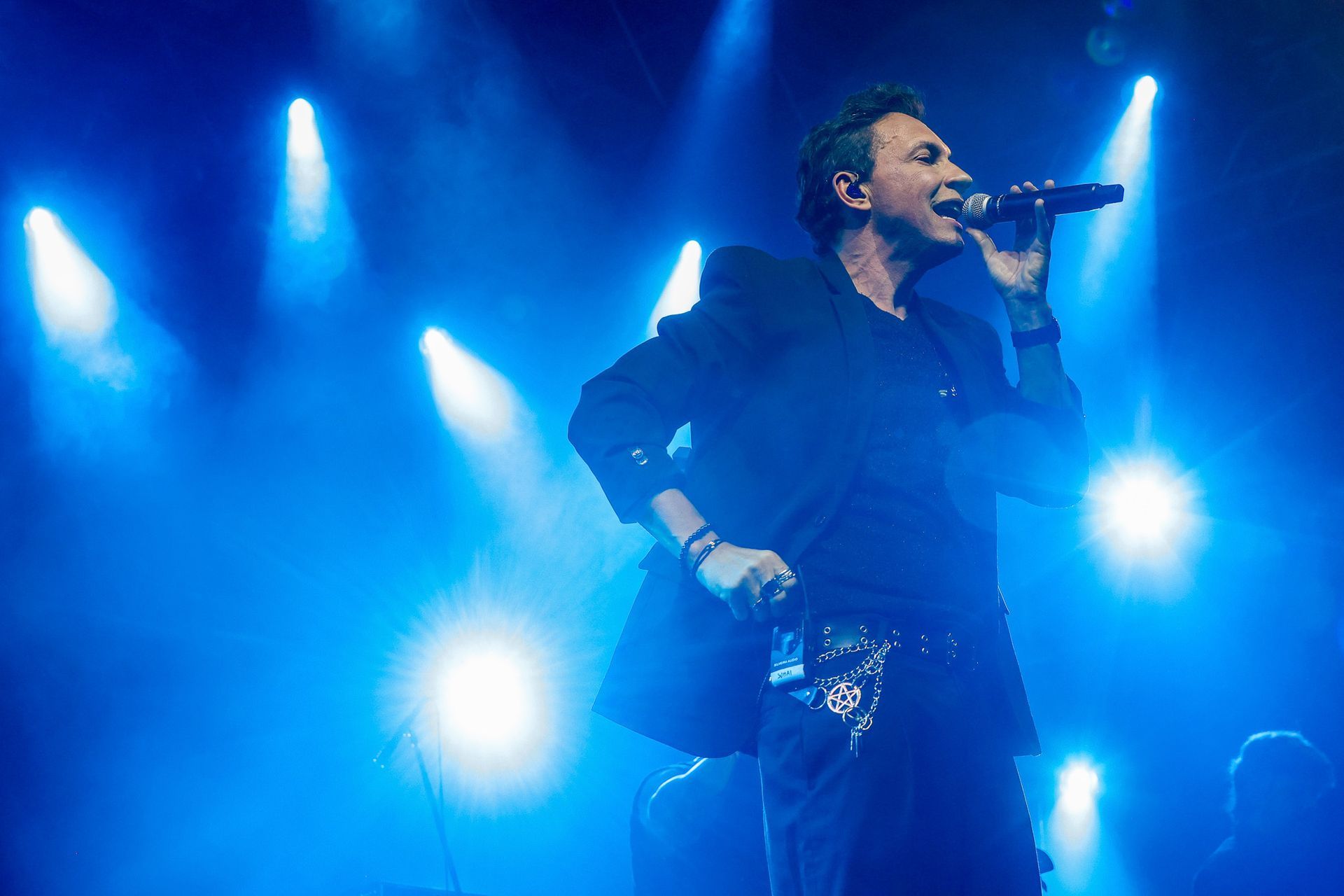 Um cantor se apresenta no palco, segurando um microfone sob holofotes azuis brilhantes em um ambiente de show.