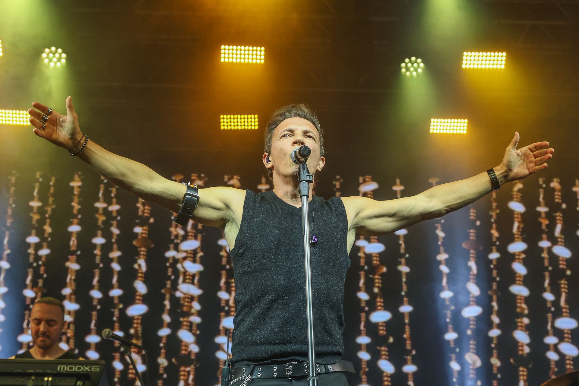 Um cantor se apresentando no palco com os braços estendidos, microfone na mão e um show de luzes brilhantes ao fundo.