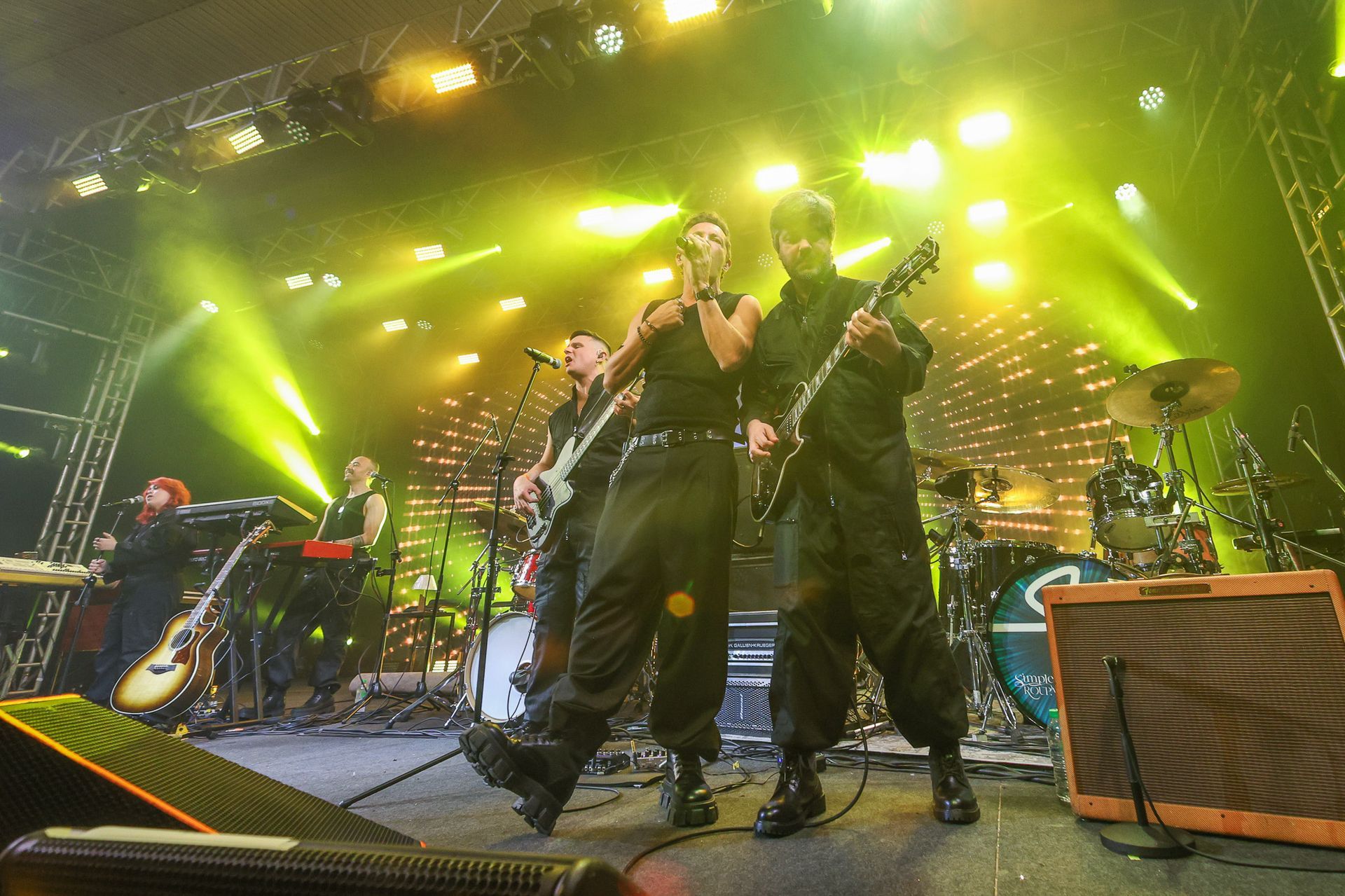 Uma banda se apresenta em um palco de shows bem iluminado com luzes amarelas, com cantores e guitarristas vestidos de preto.