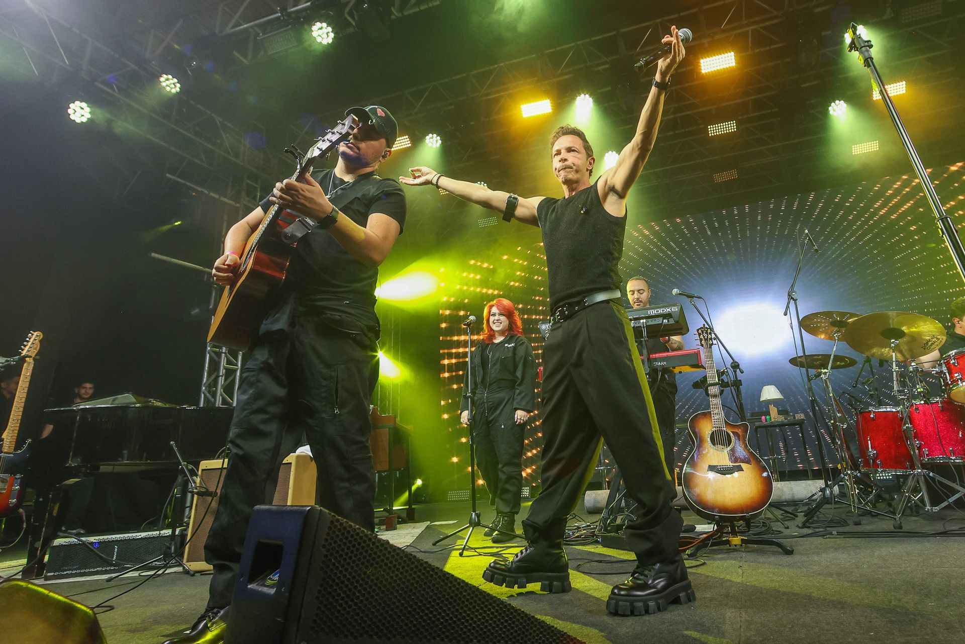 Uma banda se apresenta em um palco com iluminação amarela brilhante; o vocalista estende os braços em direção à plateia.
