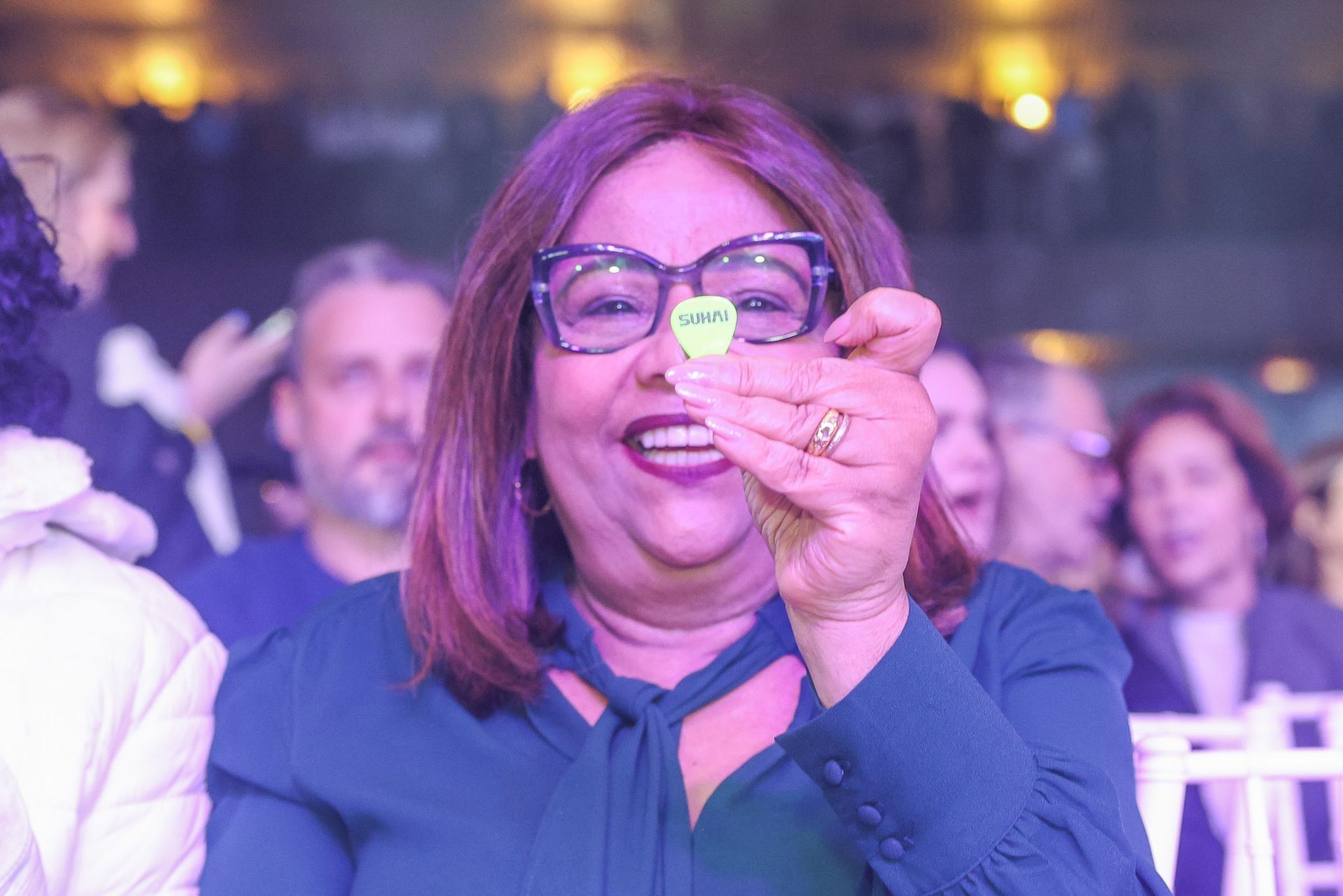Uma pessoa sorridente, vestindo uma blusa azul, segura uma pequena palheta de guitarra verde-clara em frente aos.