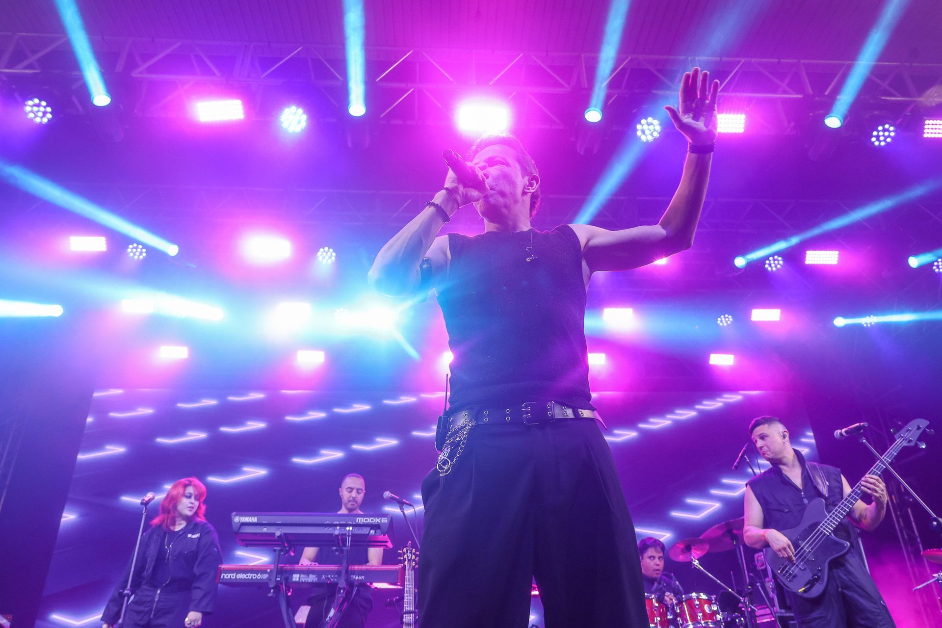Uma banda musical se apresenta em um palco de shows com iluminação vibrante em tons de rosa e azul,