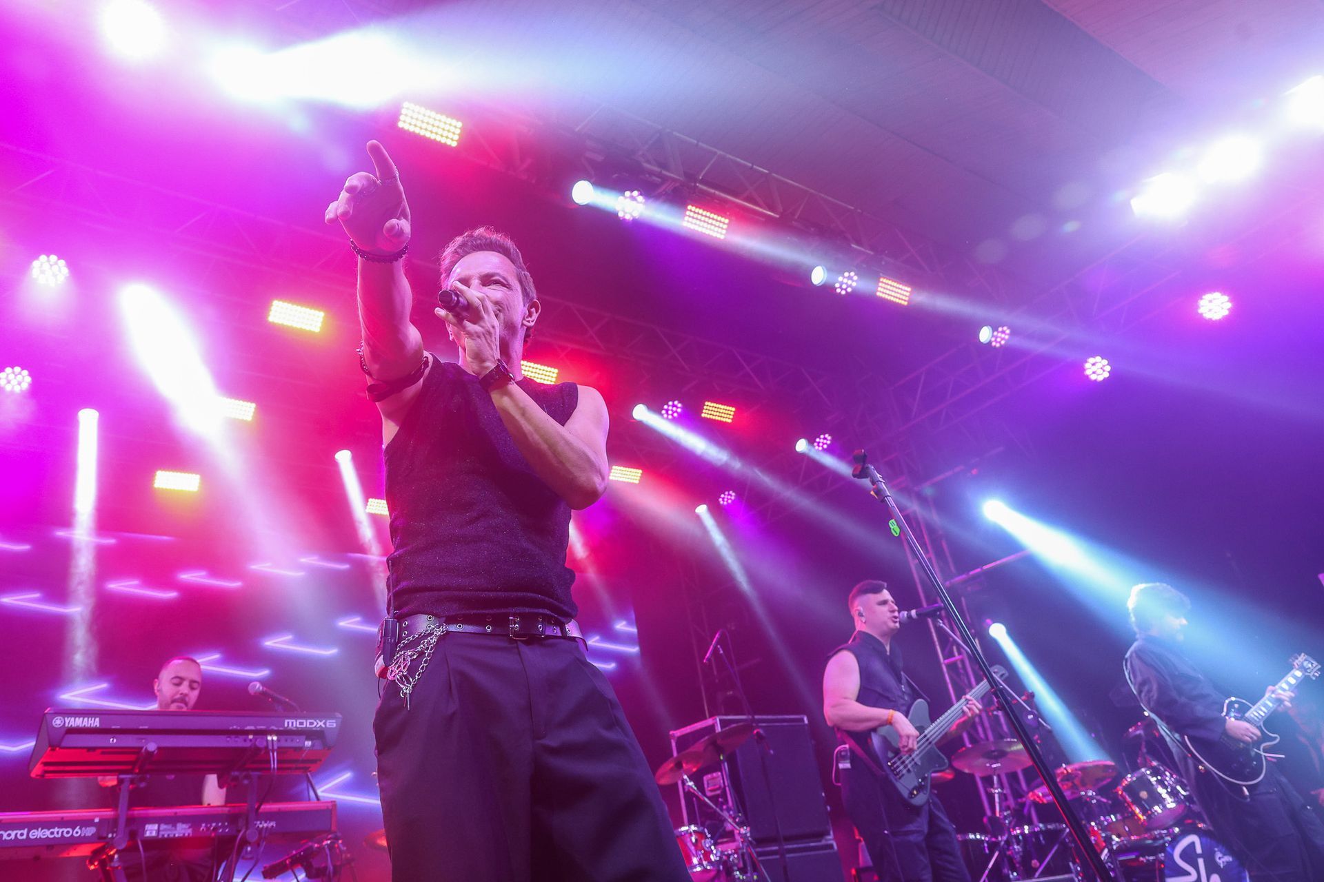 Uma banda se apresenta no palco sob holofotes roxos brilhantes, com o vocalista gesticulando em direção à plateia.