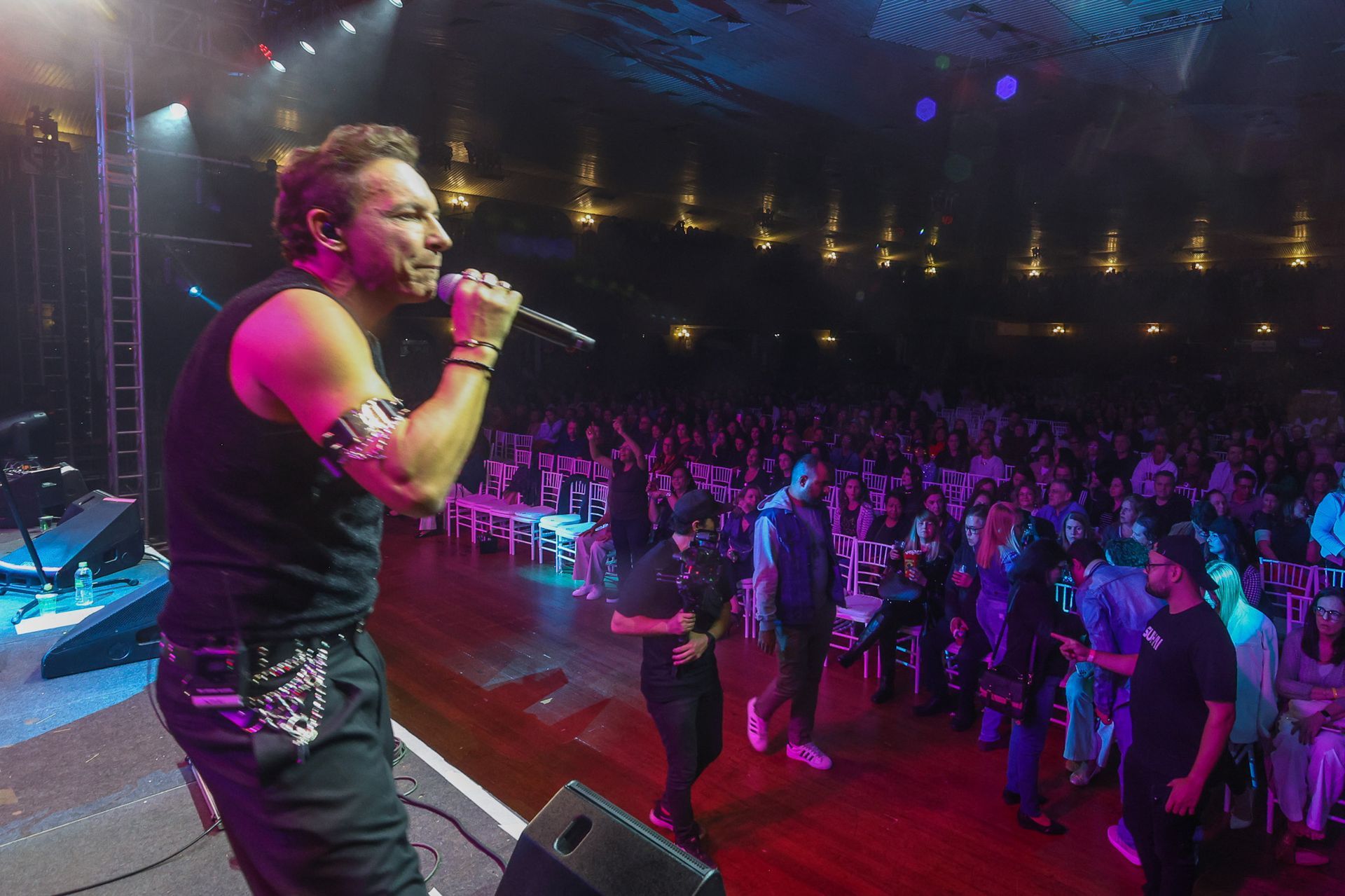Um músico de regata preta se apresenta no palco, está de frente para a plateia em uma sala de concertos com pouca luz.