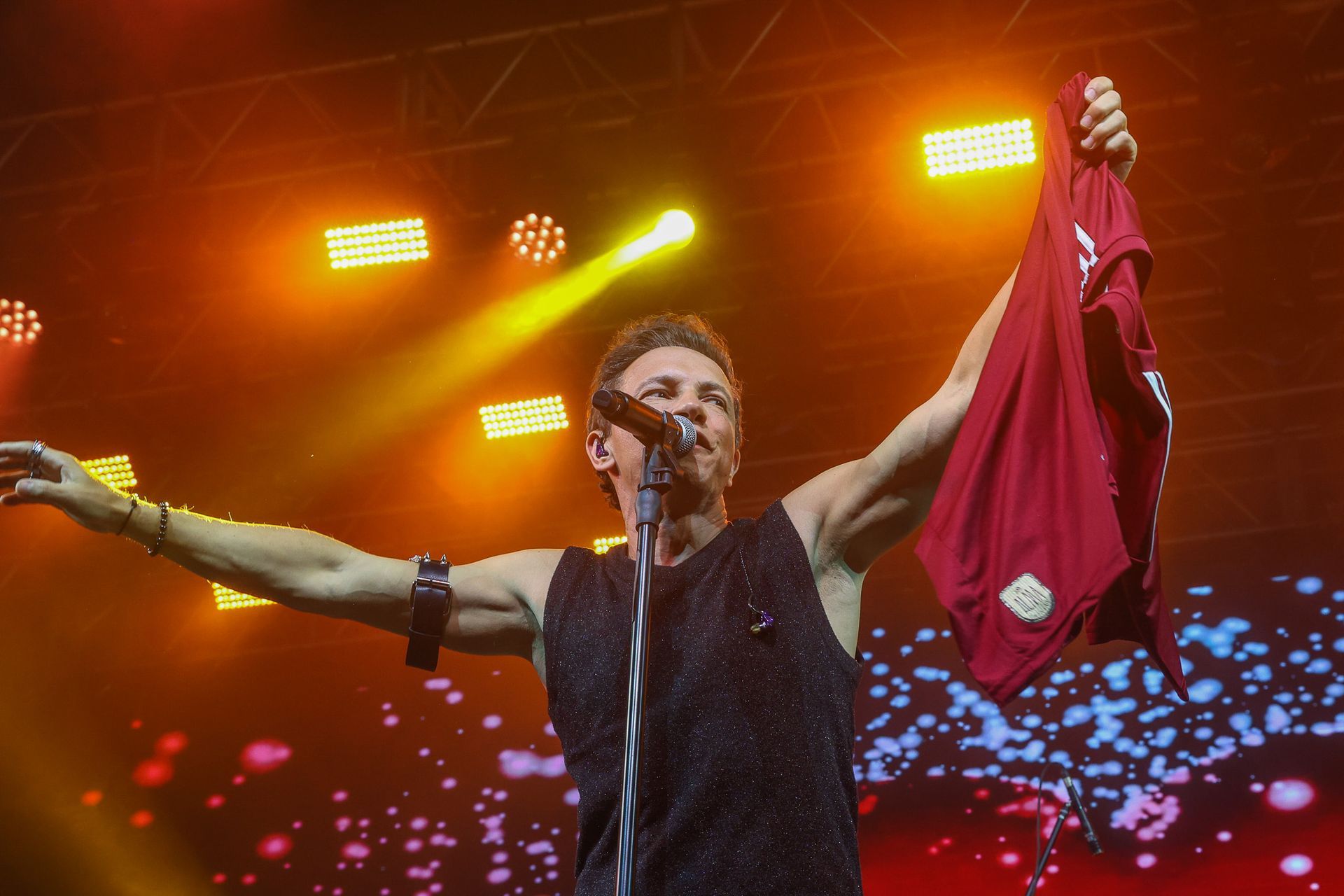 Um artista no palco segura uma peça de roupa vermelha com um braço estendido sob luzes amarelas brilhantes de um show.