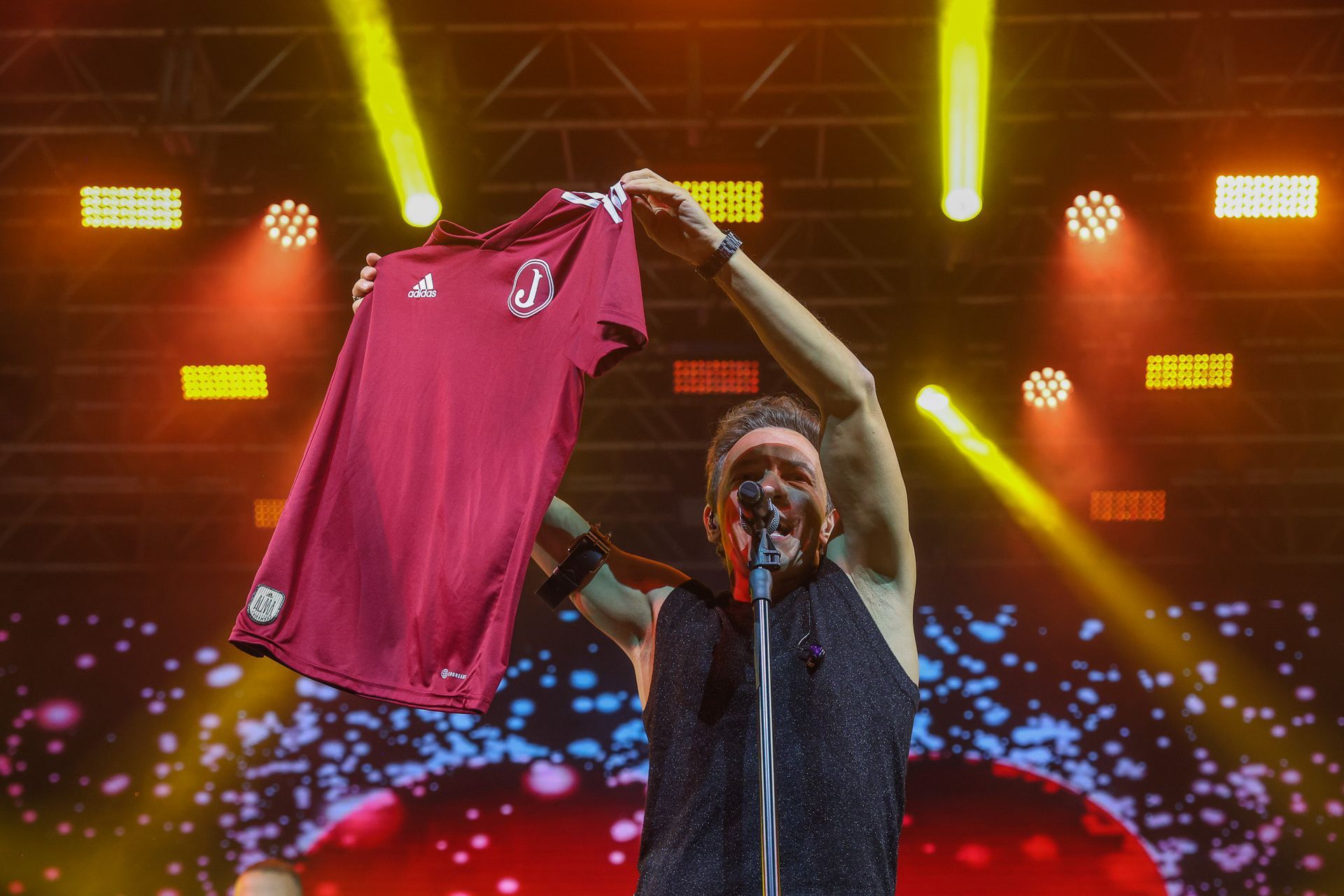 Um artista em um palco de concertos bem iluminado exibe uma camisa de futebol vermelha escura para a plateia.