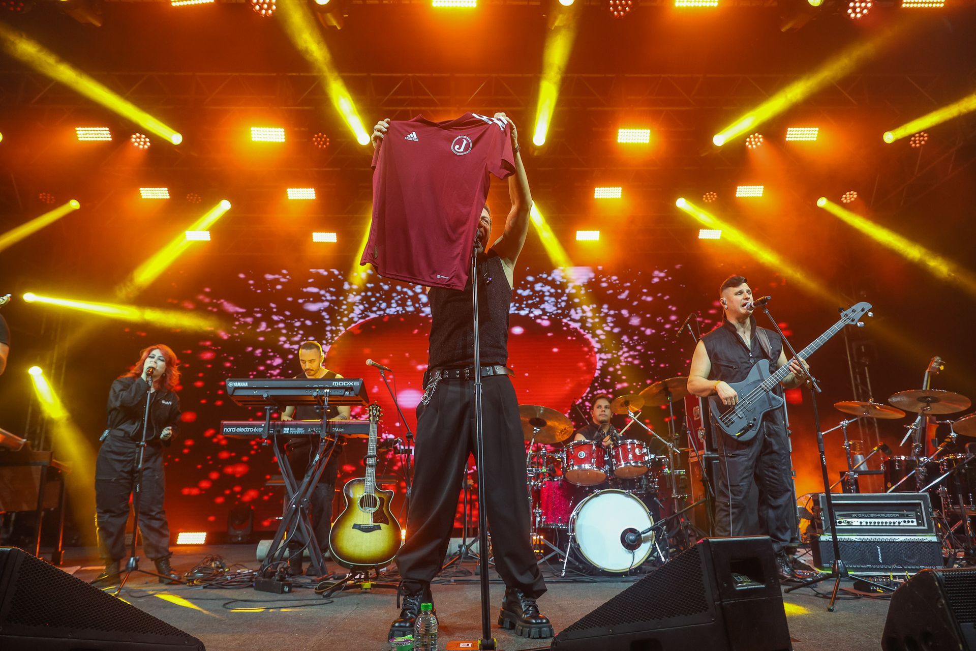 Uma banda se apresenta no palco sob holofotes brilhantes e aconchegantes; o vocalista segura uma camisa bordô
