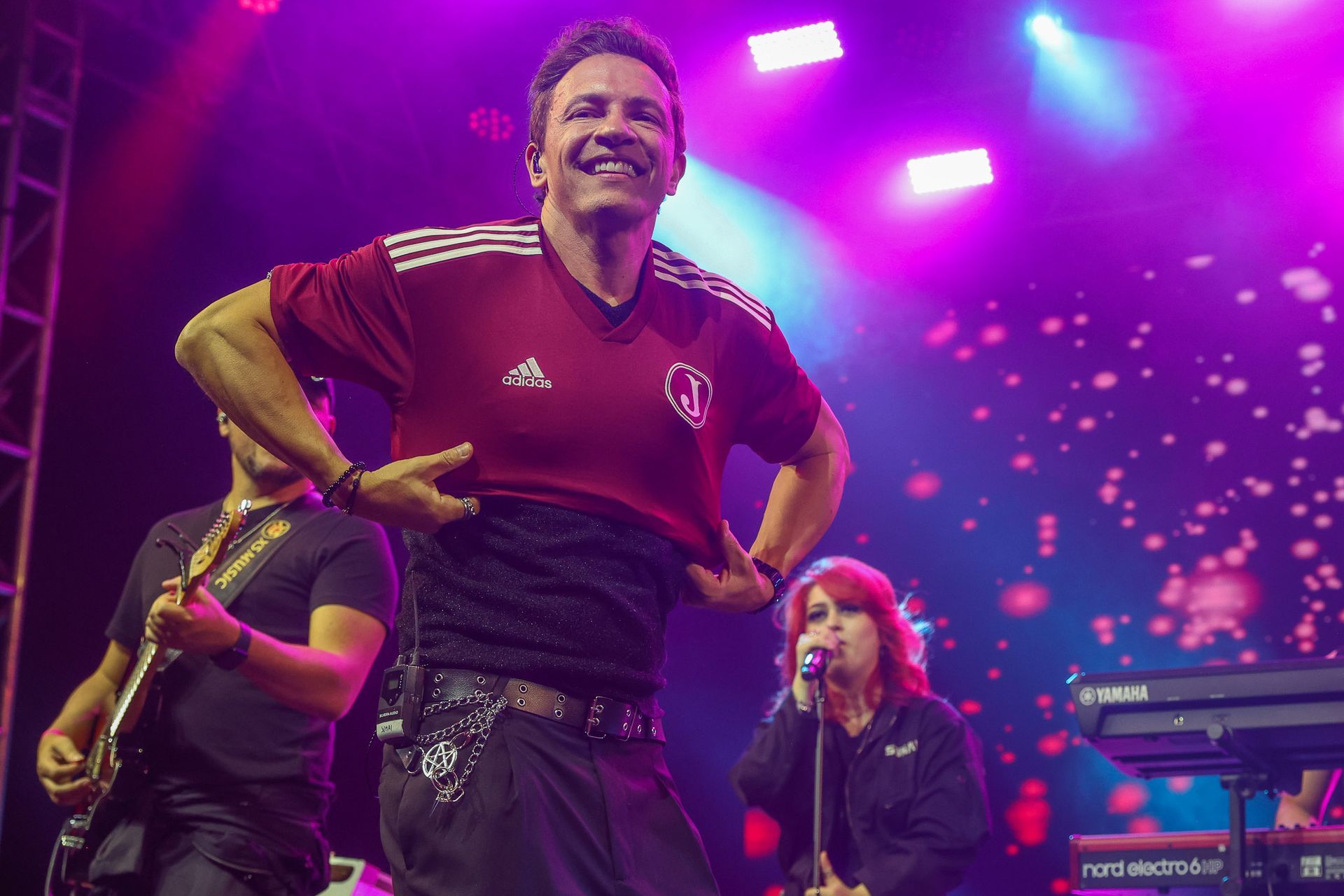 Uma pessoa no palco levanta sua camisa de futebol vinho enquanto uma banda toca ao fundo.