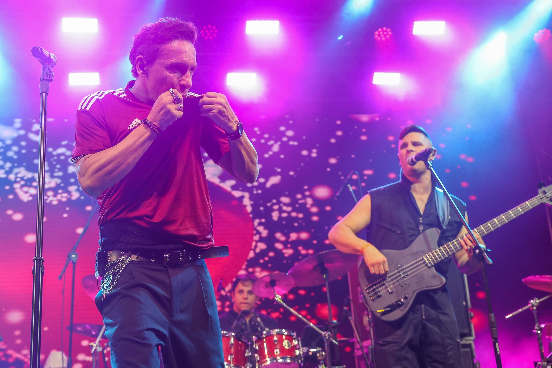 Um cantor com uma camiseta esportiva vermelha, um baixista e um baterista ao fundo.