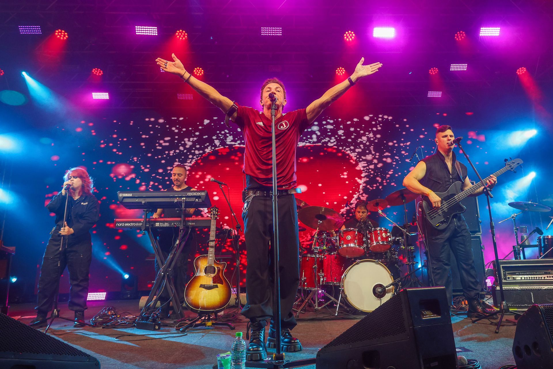 Uma banda se apresenta no palco sob luzes roxas brilhantes, com o vocalista principal em pé no centro do palco