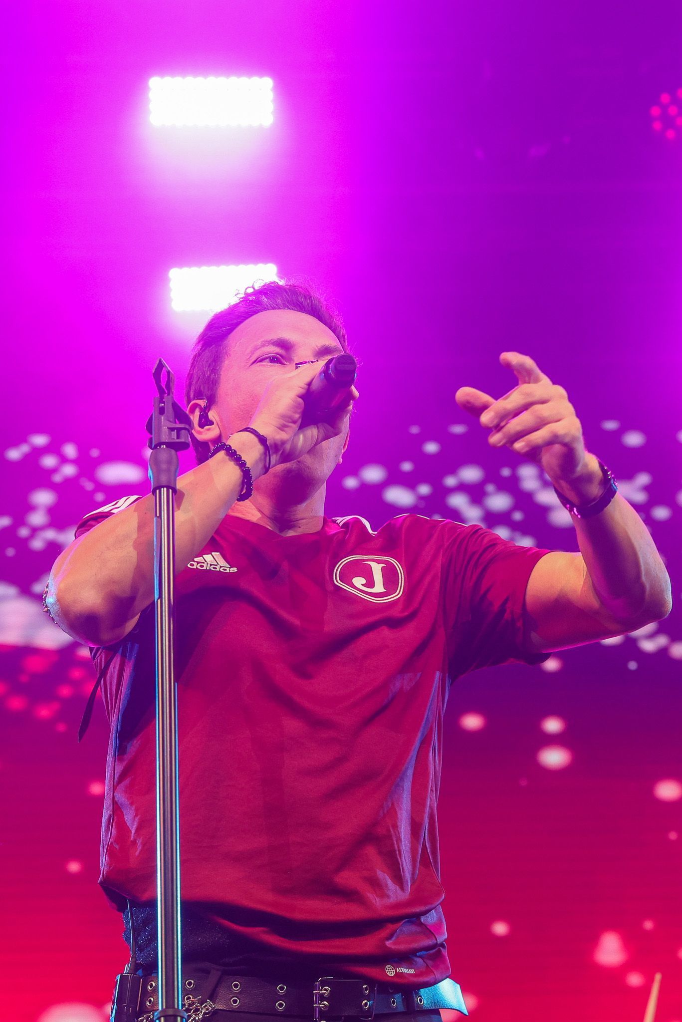 Um artista vestindo uma camisa bordô canta em um microfone no palco, iluminada por uma vibrante luz rosa.