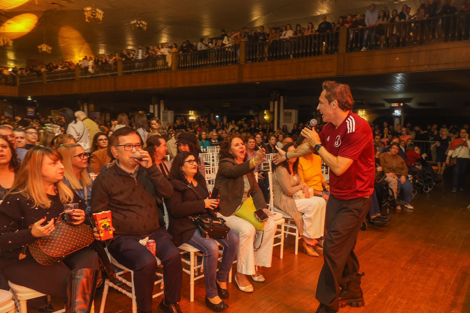 Um cantor de camisa vermelha gesticula em direção à plateia em um salão de baile pouco iluminado e com painéis de madeira.