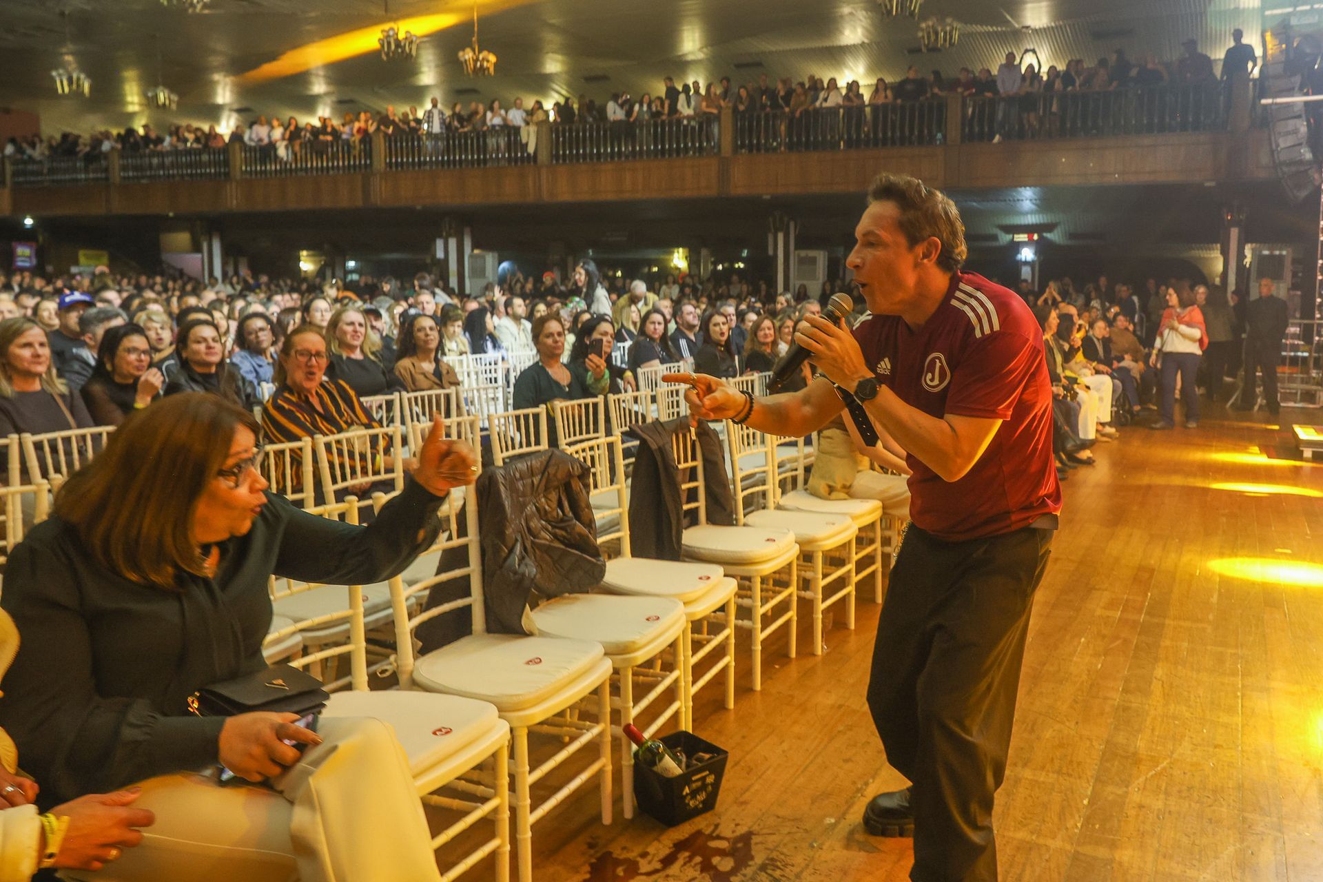 Uma cantor vestindo uma camisa esportiva vermelha fala ao microfone enquanto aponta para uma plateia
