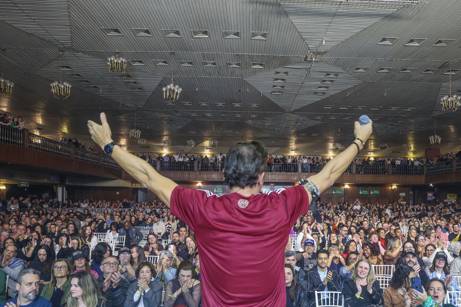 Uma pessoa vestindo uma camiseta bordô está em pé no palco com os braços erguidos diante de uma grande plateia