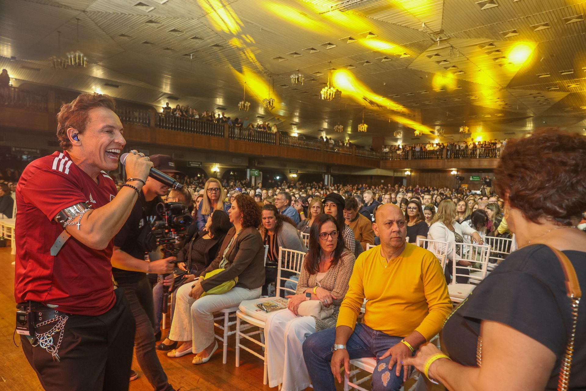 Um cantor vestindo uma camisa vermelha se apresenta com um microfone diante de uma plateia em uma grande sala de concertos