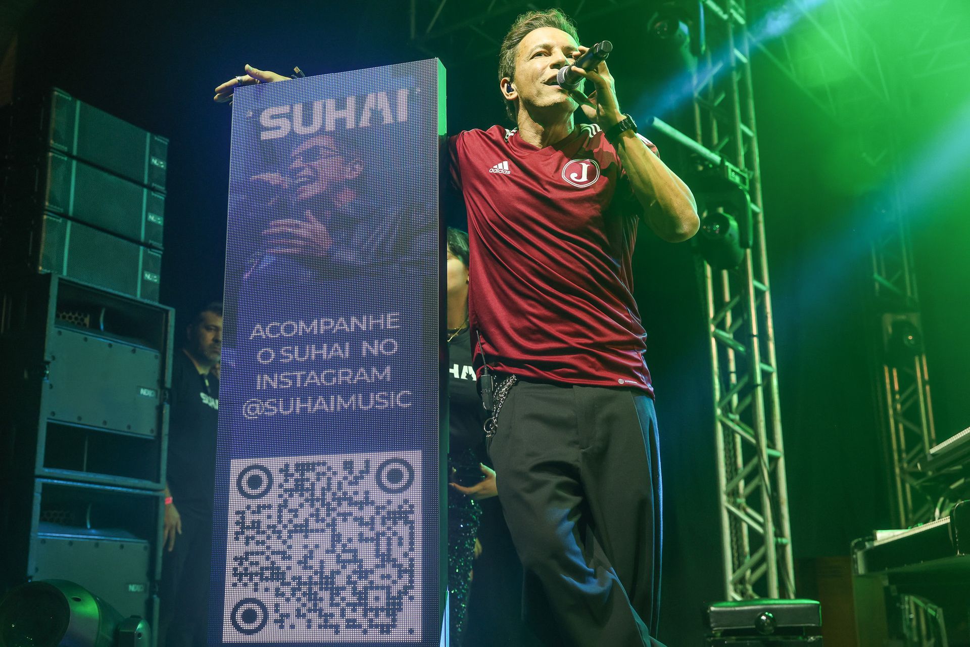 Uma pessoa vestindo uma camisa bordô cantando em um microfone no palco, ao lado de um painel alto e iluminado 