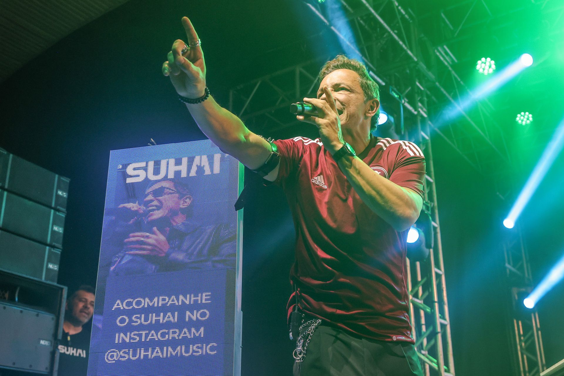 Uma pessoa vestindo uma camisa bordô cantando em um microfone no palco, ao lado de um painel alto e iluminado 