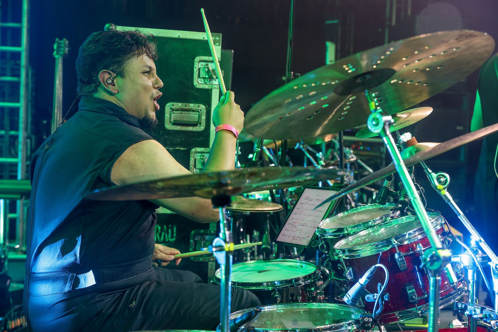 Um baterista tocando bateria no palco, concentrado com a boca aberta, cercado por pratos, tambores e iluminação cênica.
