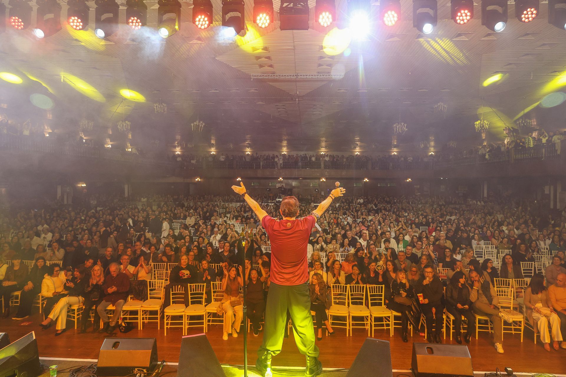 Um artista vestindo camisa vermelha está em pé no palco com os braços erguidos diante de uma grande plateia