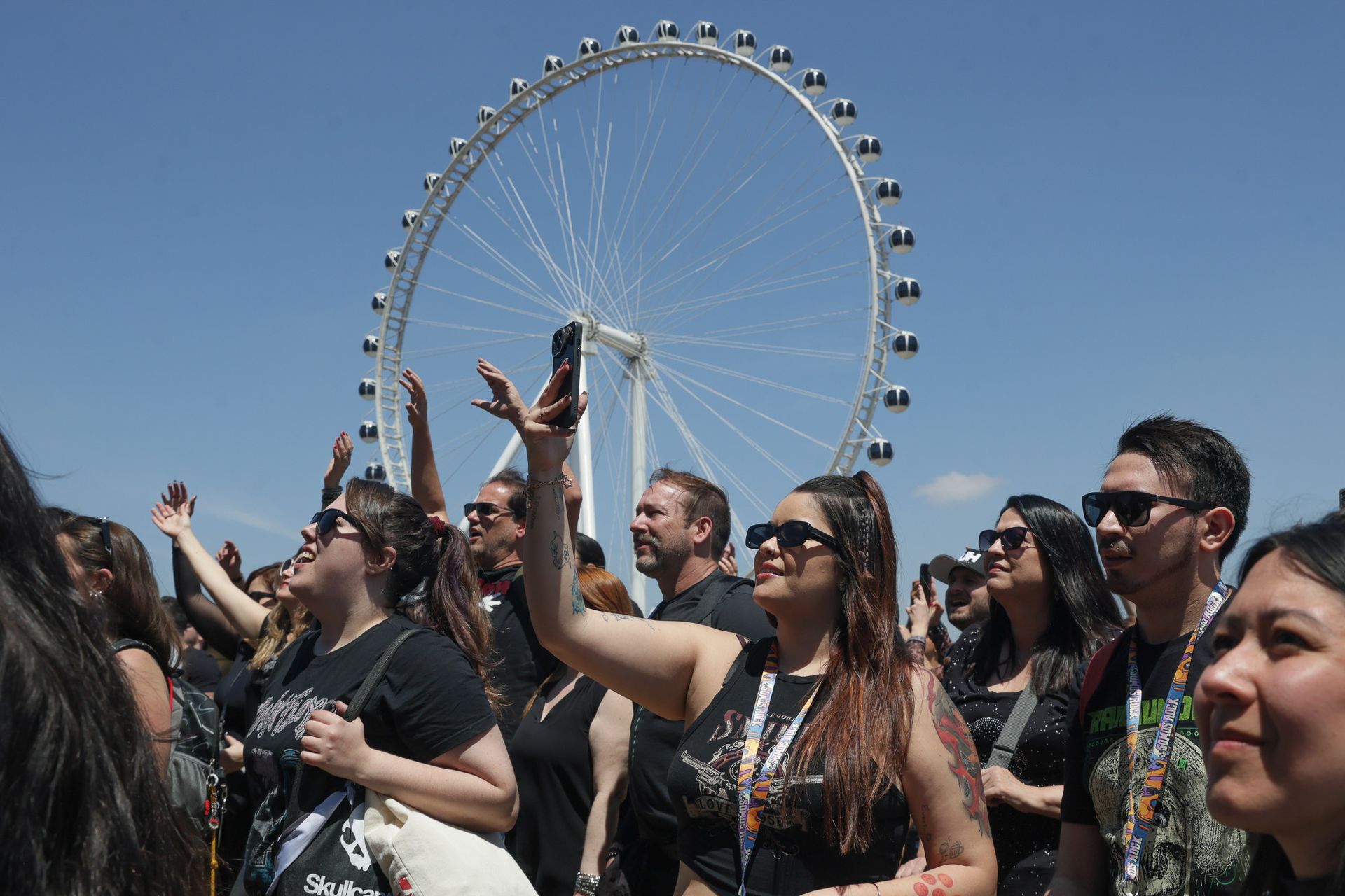 Uma multidão ao ar livre em um festival ensolarado, com pessoas tirando fotos, em frente a uma grande roda-gigante.