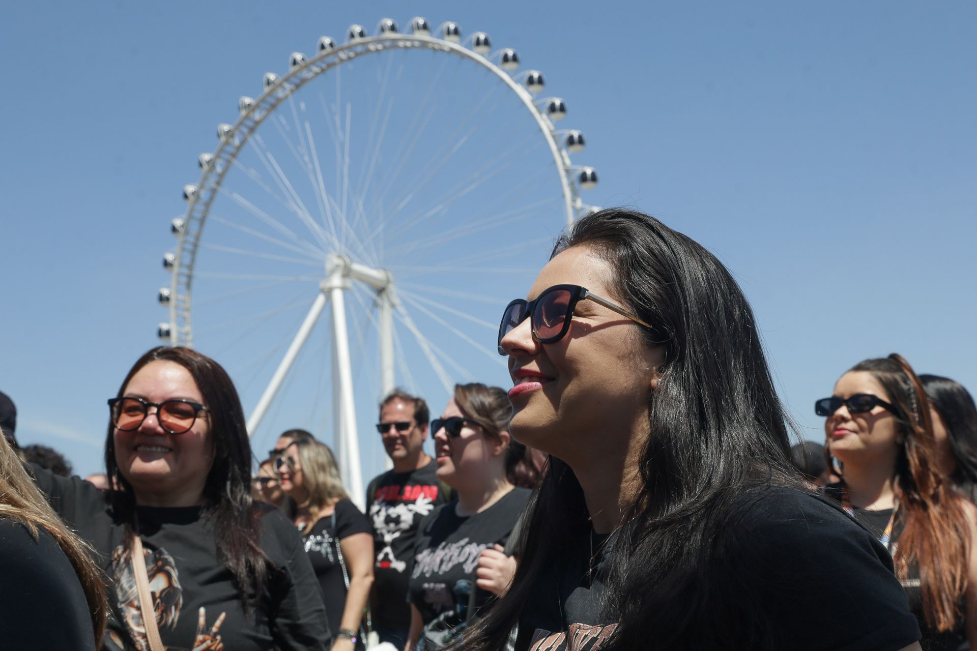 Um grupo de pessoas vestindo camisas pretas sorri ao ar livre em frente a uma grande roda-gigante sob um céu azul claro.