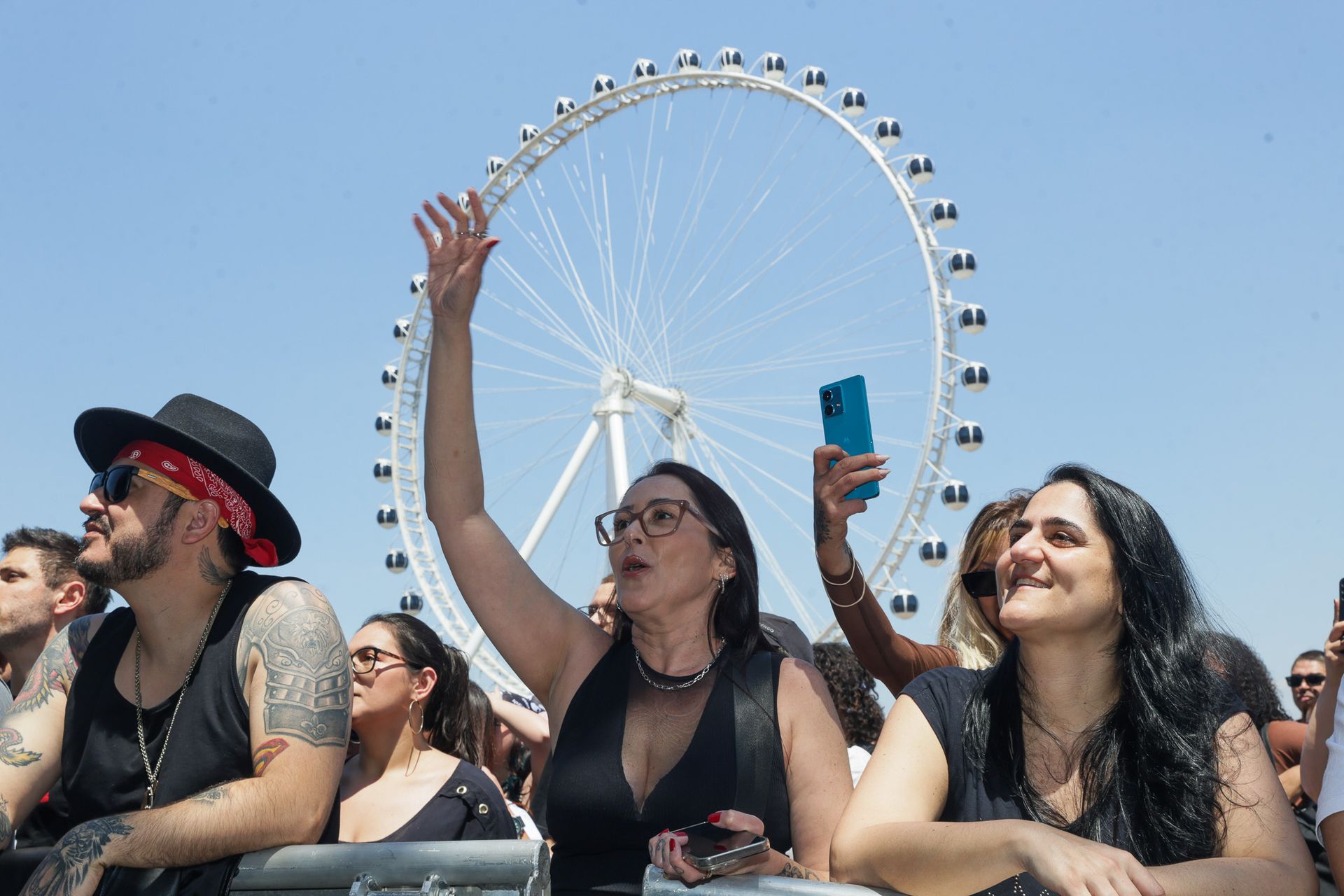 Uma multidão em um evento ao ar livre com uma grande roda-gigante ao fundo; uma pessoa com o braço levantado.