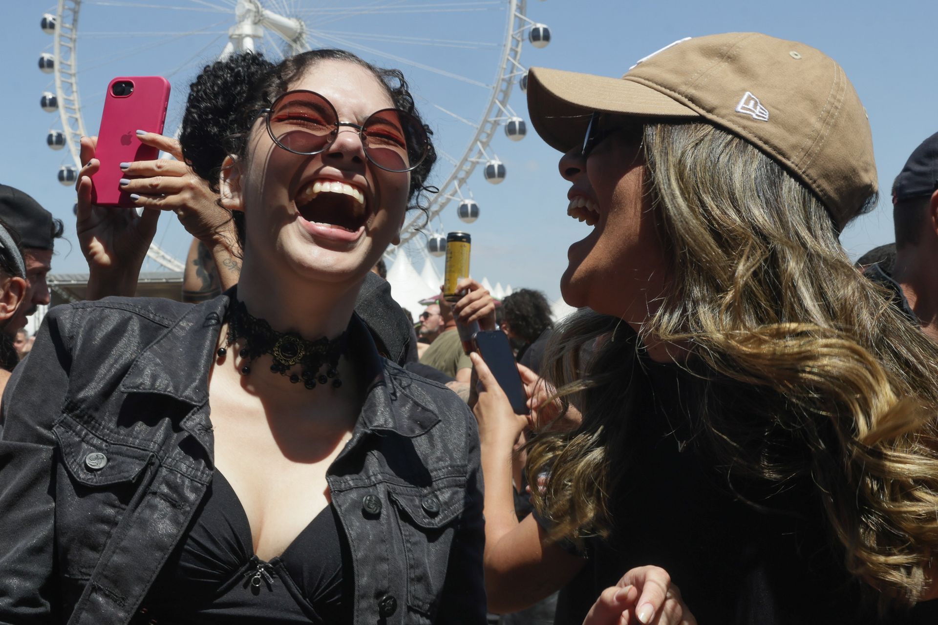 Duas pessoas rindo ao ar livre com uma roda-gigante ao fundo, uma delas segurando um celular rosa.