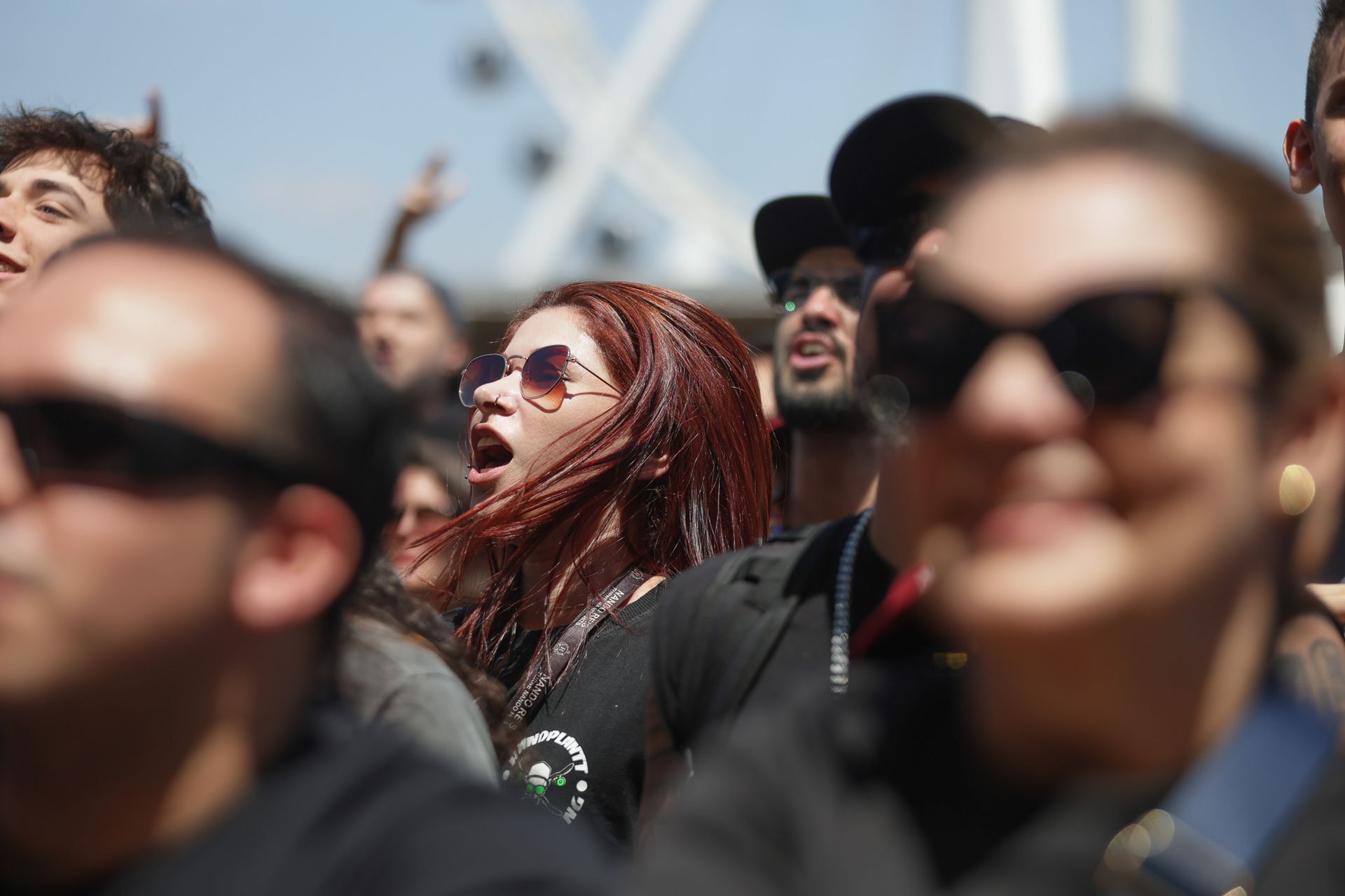 Uma multidão de pessoas usando óculos escuros em um evento ao ar livre, com uma mulher no centro cantando de boca aberta.