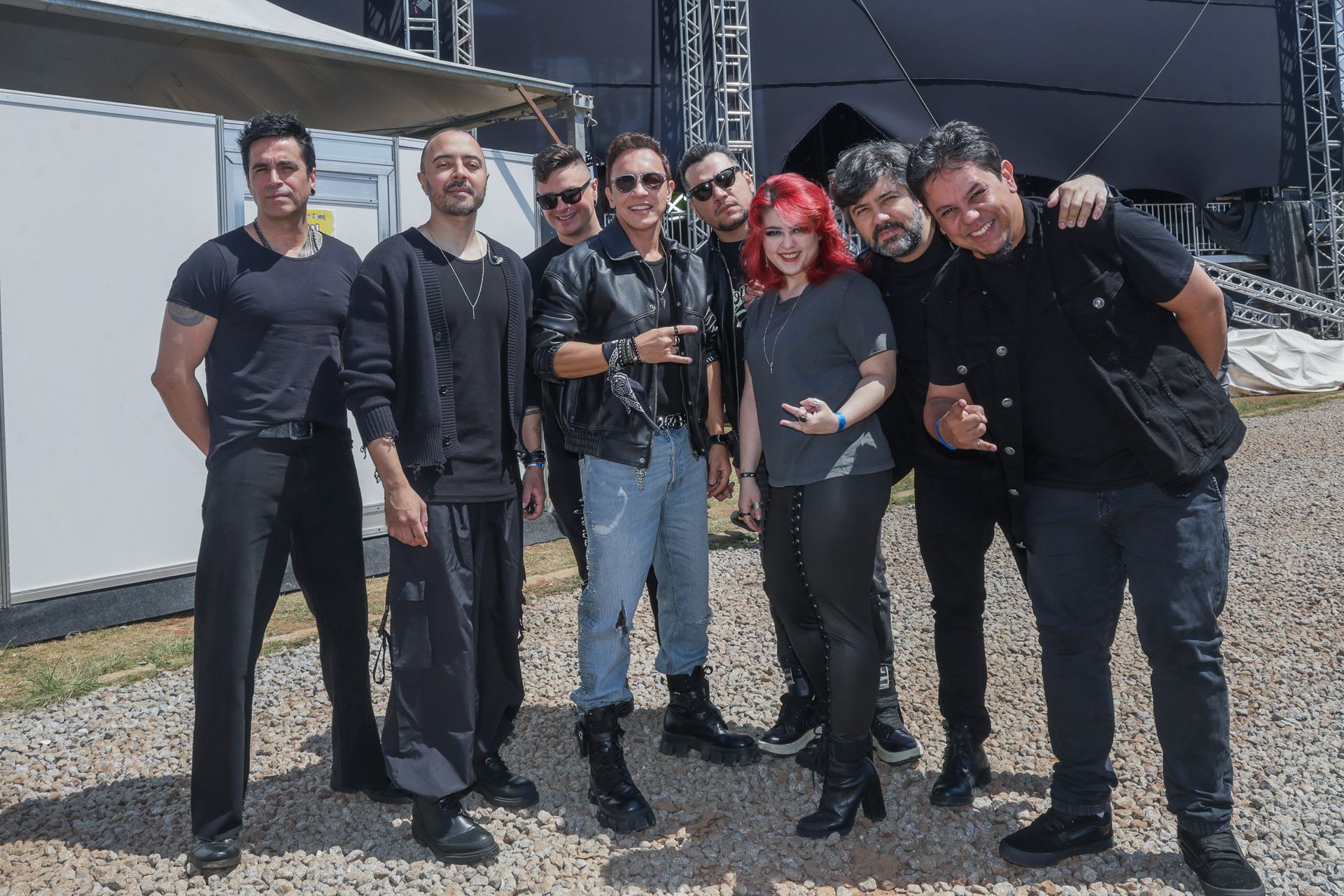 Um grupo de oito pessoas posando juntas ao ar livre em frente a um palco, vestindo roupas casuais em tons escuros.