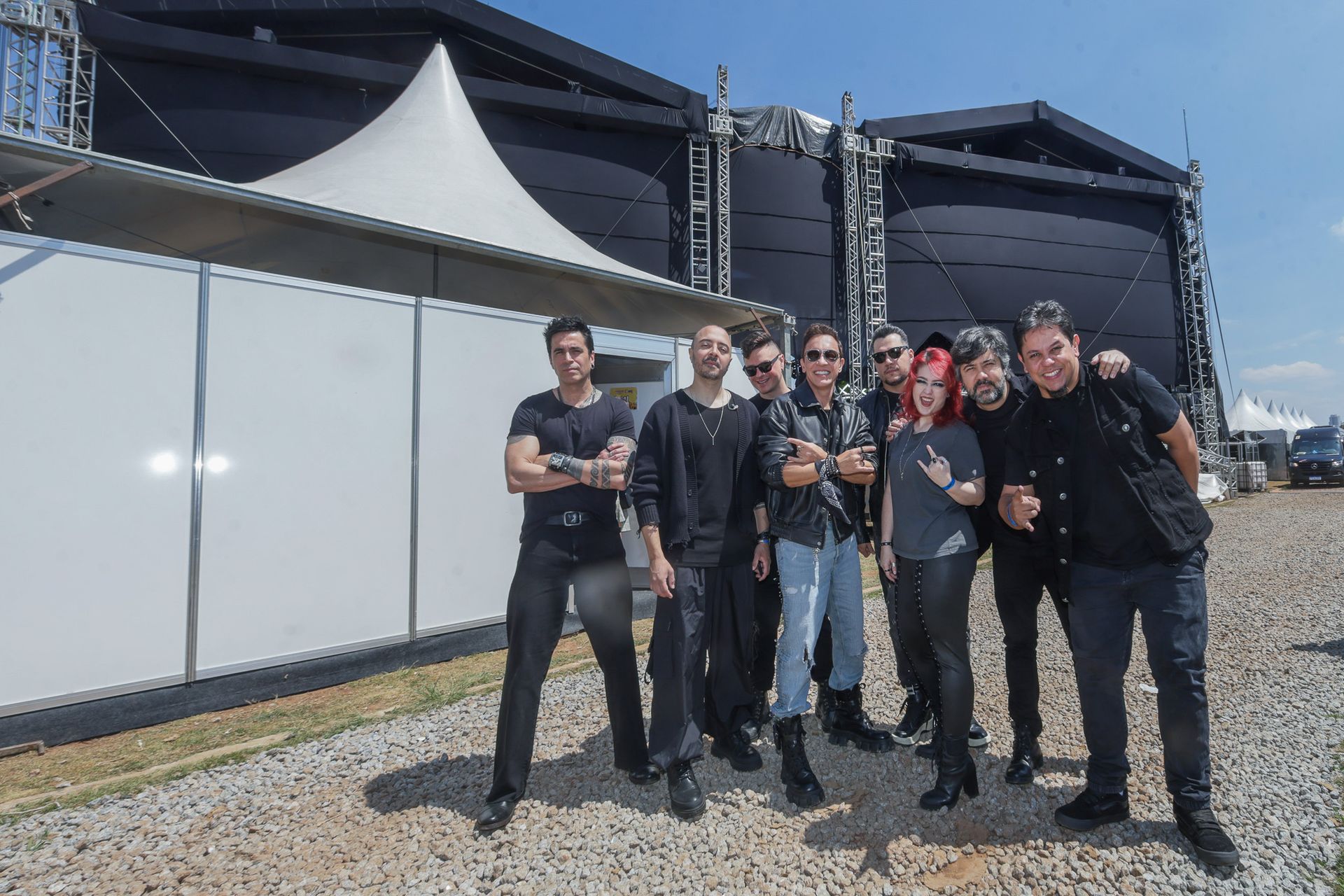 Um grupo de oito pessoas vestindo roupas escuras posa junto em frente a uma grande estrutura coberta por uma tenda.