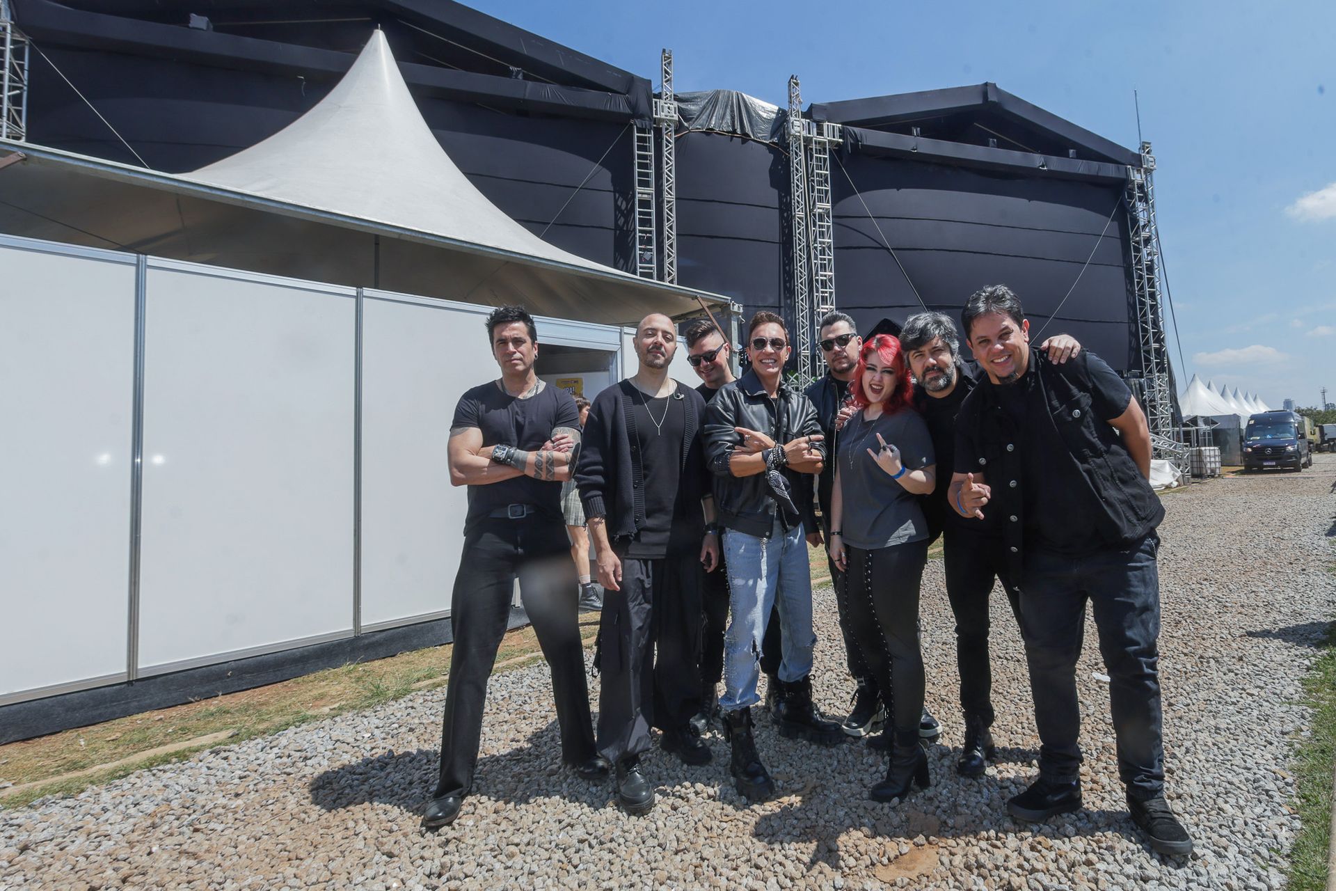 Um grupo de oito pessoas vestindo roupas casuais escuras posando juntas  perto de uma tenda de palco sob um céu azul.