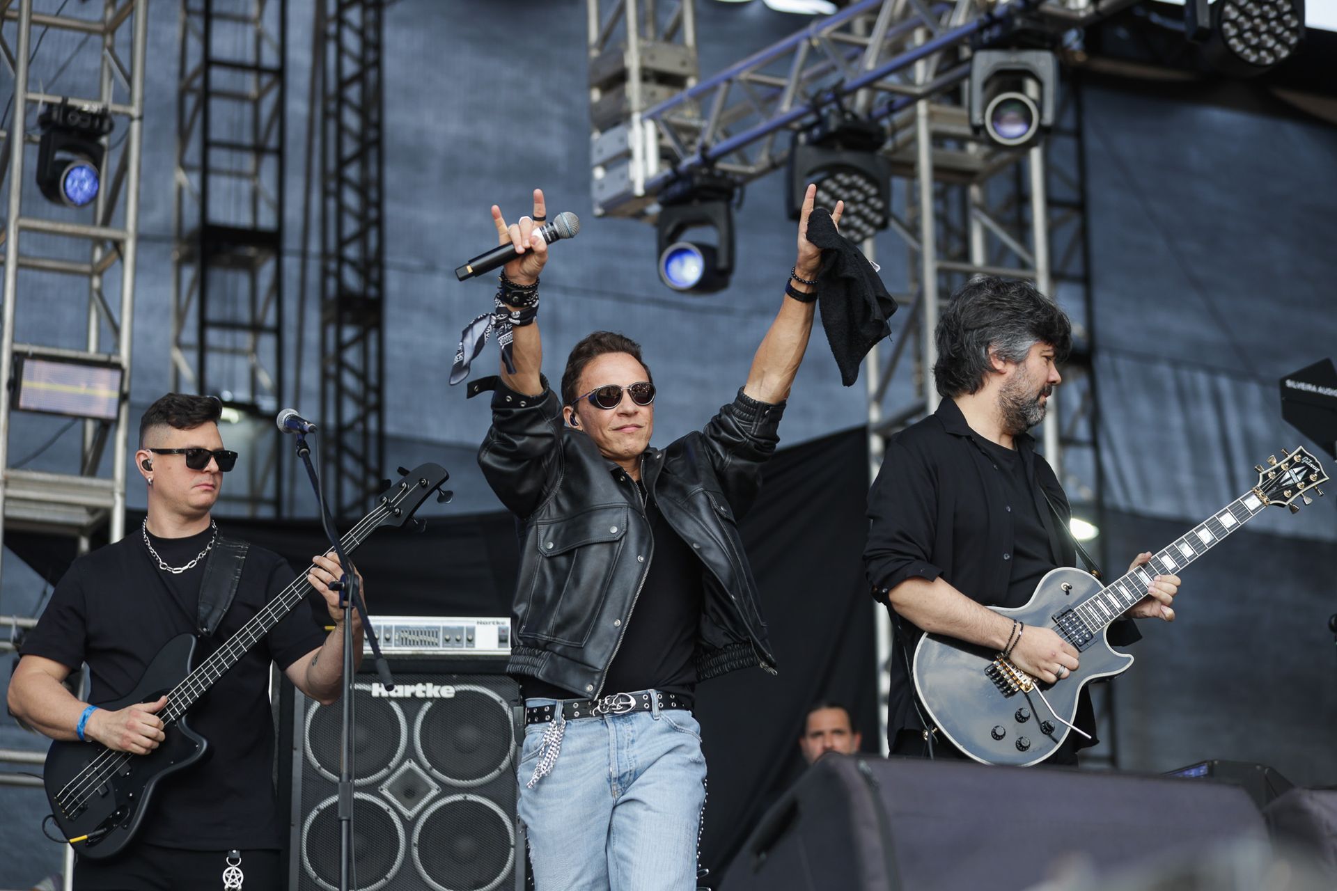 Uma banda em um palco de shows, com um vocalista central acenando e dois instrumentistas tocando guitarra e baixo.