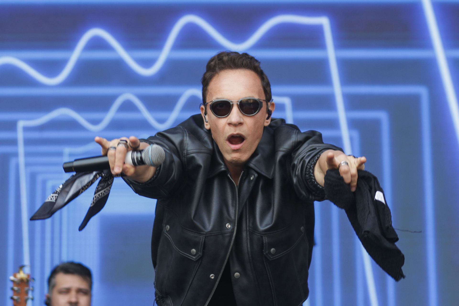 Um artista vestindo jaqueta de couro preta e óculos escuros inclina-se para a frente um fundo azul com detalhes em neon.