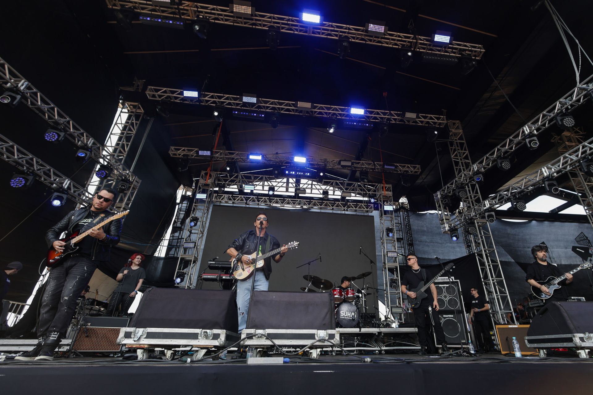 Uma banda se apresenta em um palco de shows com equipamentos de iluminação, amplificadores e telões ao fundo.