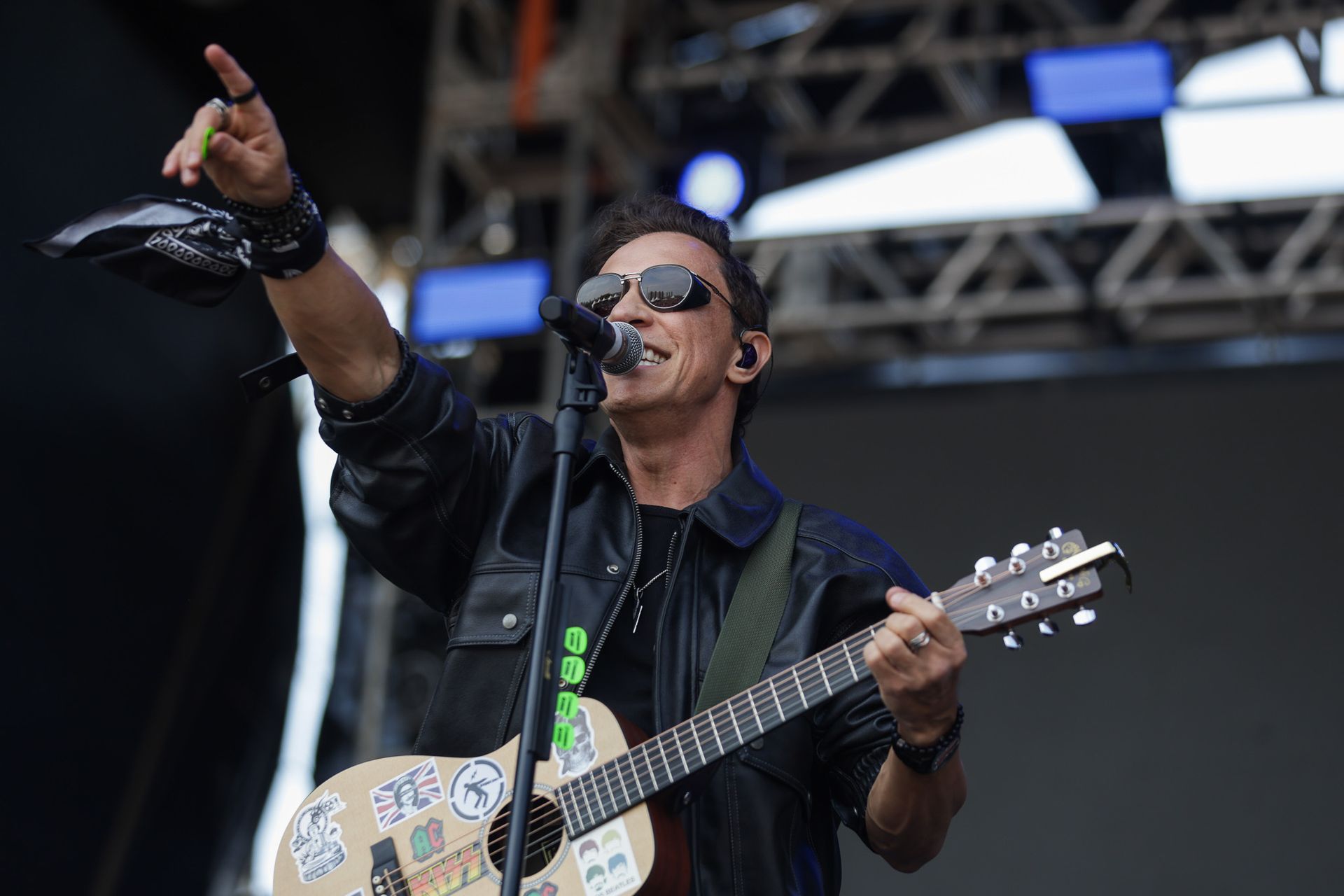 Um músico de óculos escuros e jaqueta de couro preta toca um violão coberto de adesivos enquanto aponta para o palco.