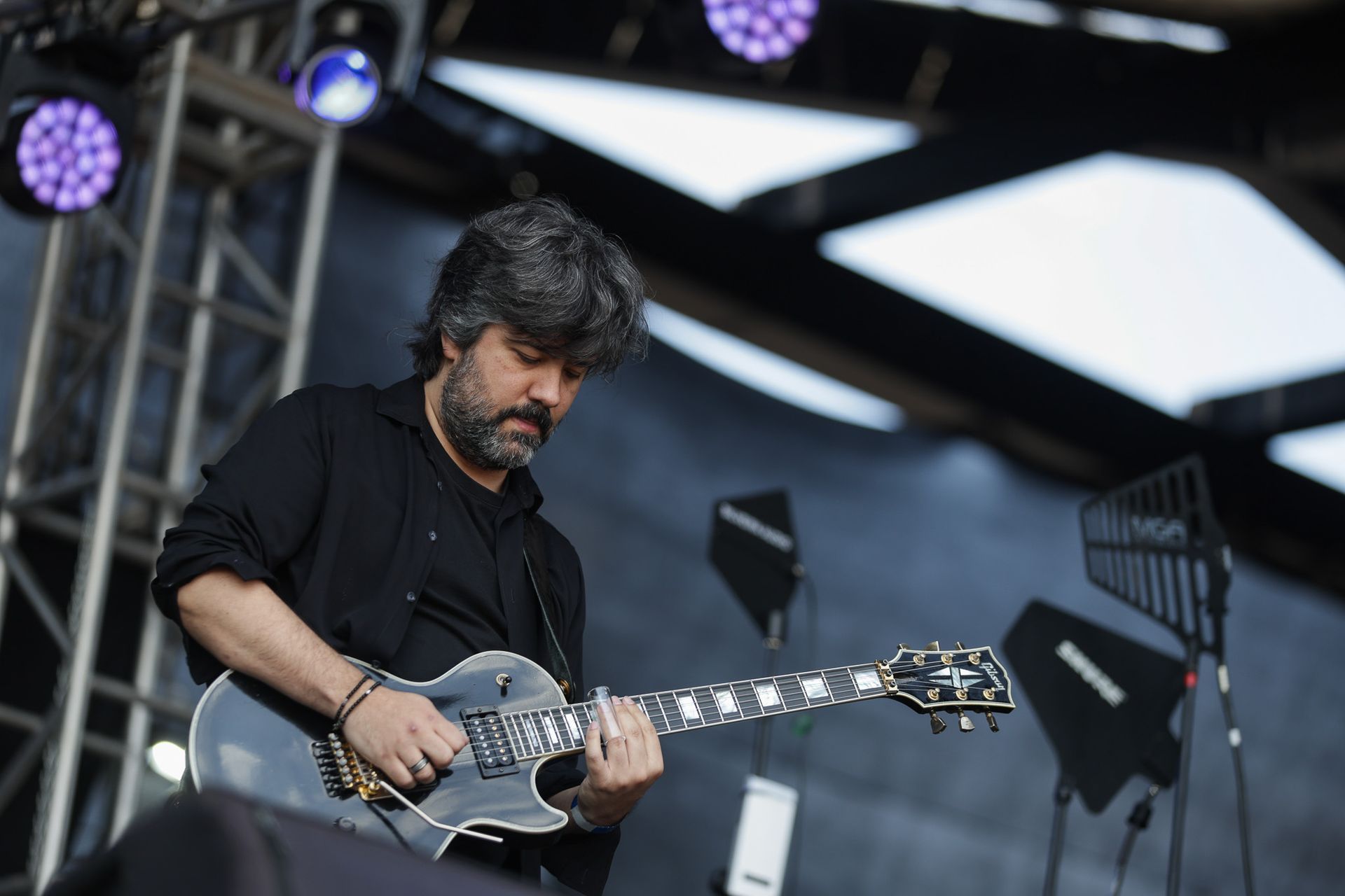 Uma pessoa de cabelos escuros e barba tocando uma guitarra elétrica preta no palco sob luzes de concerto.