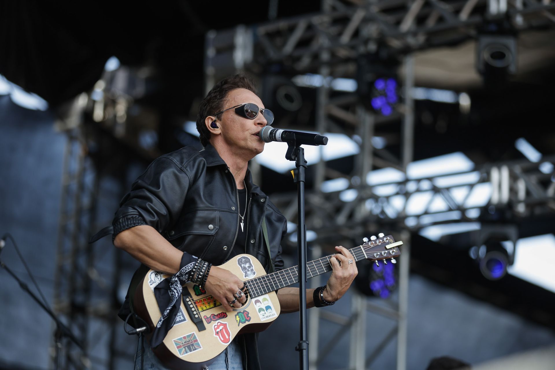 Um músico usando óculos escuros e jaqueta de couro toca um violão coberto de adesivos enquanto canta em um microfone.