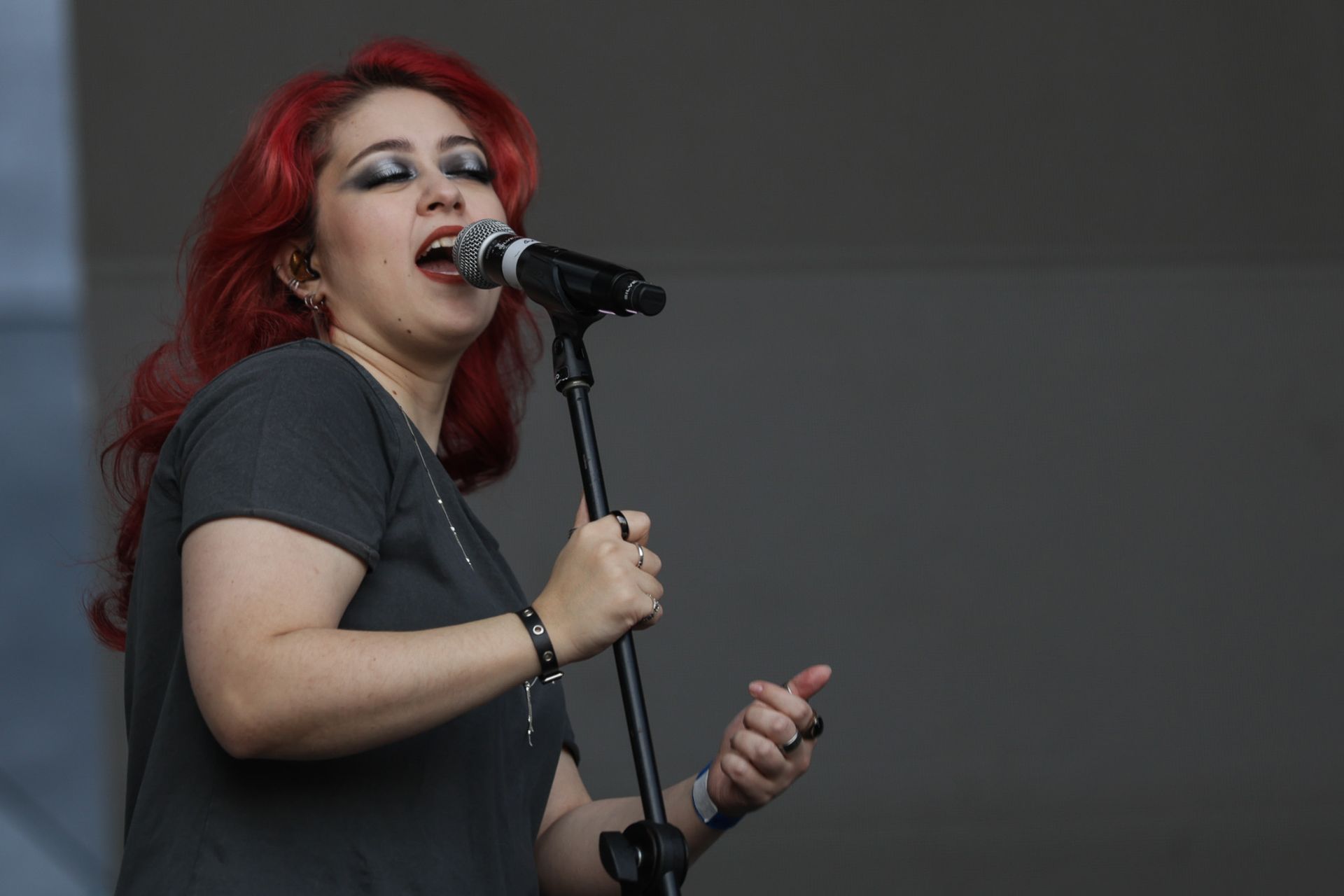 Uma pessoa com cabelos ruivos vibrantes, vestindo uma camiseta cinza, cantando com paixão em um microfone no palco.