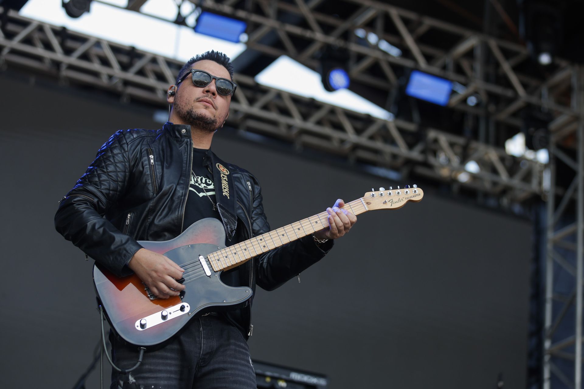 Um músico com jaqueta de couro preta e óculos escuros toca uma guitarra com em um palco de concerto.