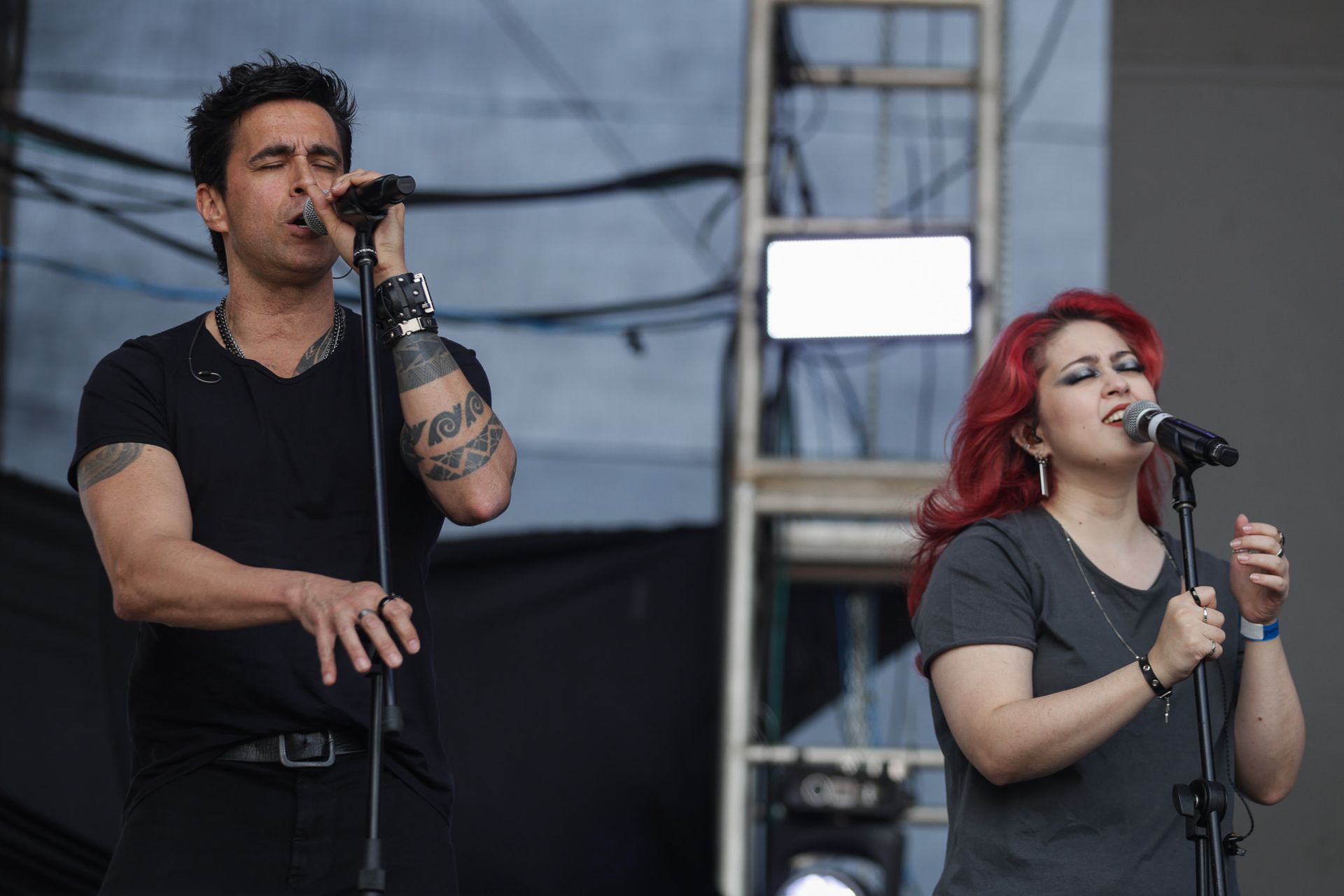 Um artista de camisa preta e uma mulher ruiva cantando em microfones em um palco ao ar livre.