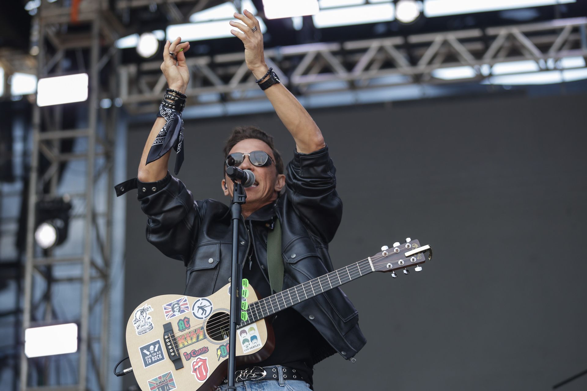 Uma pessoa vestindo jaqueta de couro e óculos escuros toca violão com adesivos no palco, com as mãos erguidas.