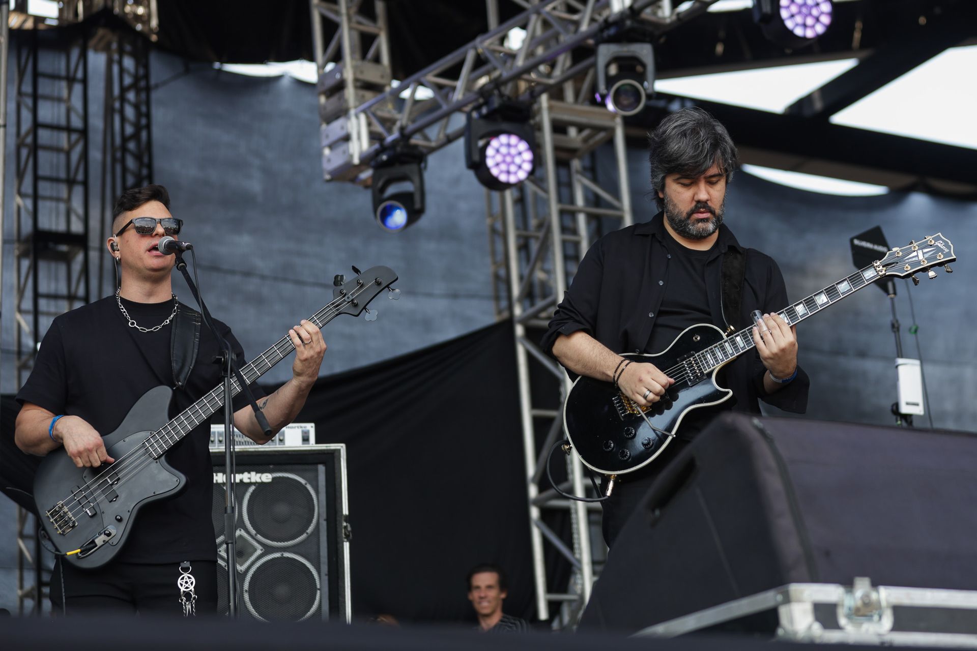 Dois músicos se apresentam no palco com guitarras, tendo um fundo escuro e uma estrutura de iluminação cênica.